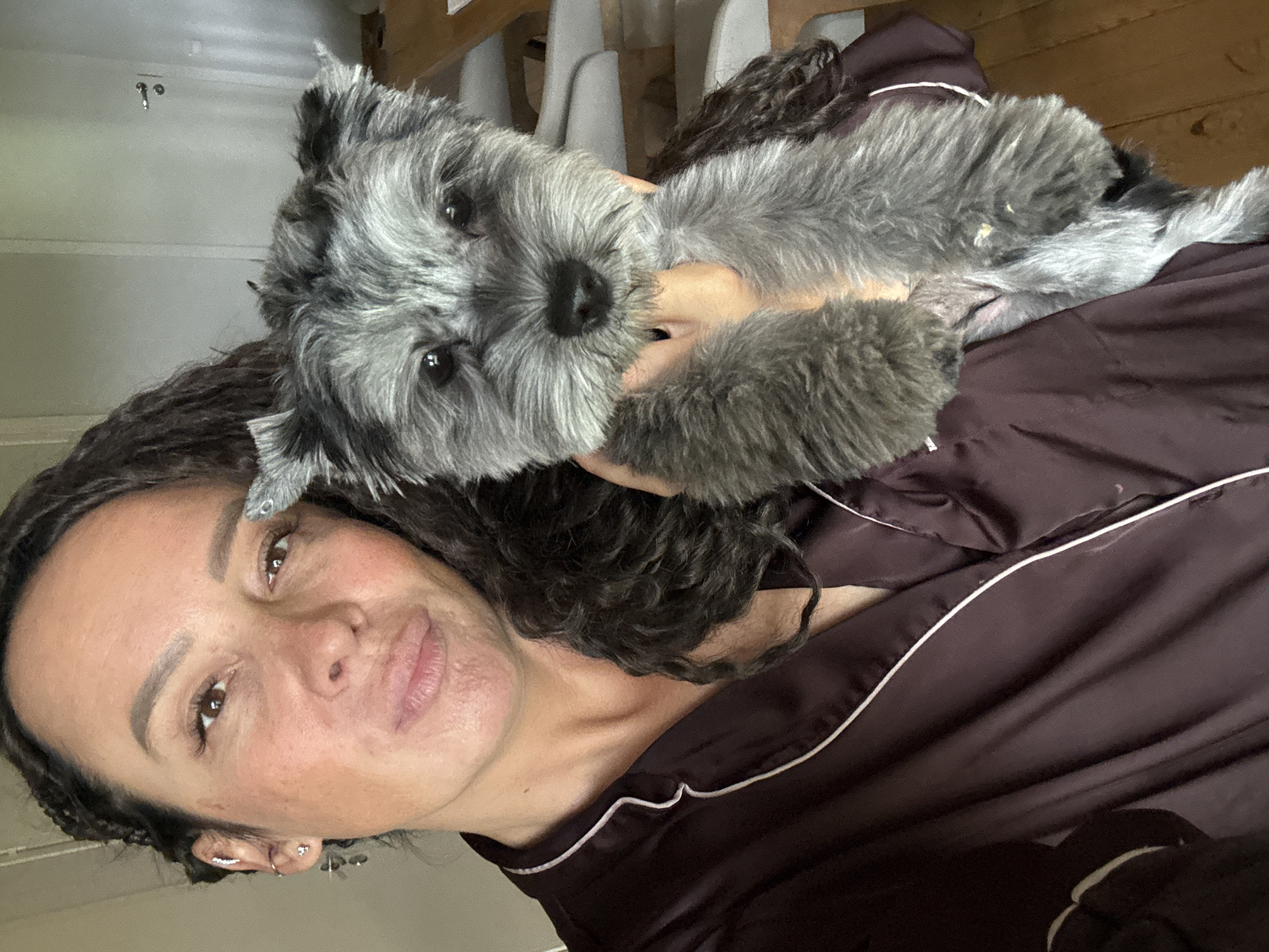 The image features a woman holding a small, grey and black terrier-type dog indoors. The woman, positioned on the left side of the frame, has dark, curly hair, brown eyes, and a fair complexion with visible freckles. She wears several earrings on her visible left ear and a dark brown robe with white piping. Her lips are slightly pursed as she looks directly at the camera. The dog, held in her right arm, has predominantly grey, shaggy fur with darker grey or black markings around its muzzle and ears. It has dark eyes, a black nose, and is also looking directly at the camera. The background consists of light-colored walls, a white door with a silver handle on the right side of the frame, and a light wooden floor visible in the lower right. Blurred, light-colored furniture is also visible in the mid-right background. There are no discernible visual cues within the image to indicate its specific location in the city of Chaikovsky, Russia, or any other specific geographical location.