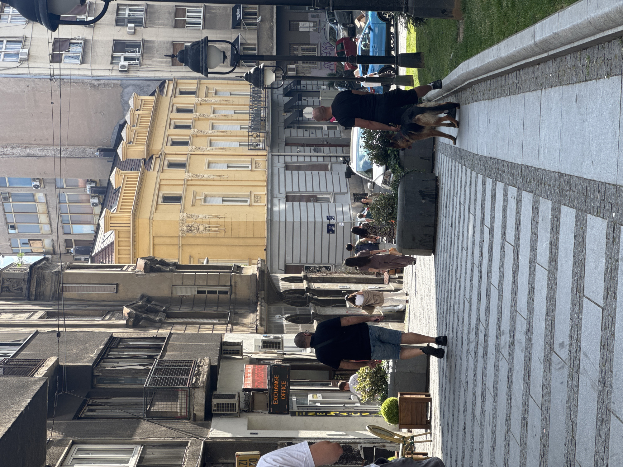 The image captures a street scene in Belgrade, Serbia, characterized by a mix of old and new architecture under a bright, sunny sky. The perspective is from ground level, looking down a pedestrian walkway paved with alternating bands of grey stone and darker, textured stone.
In the foreground, a bald man wearing a black t-shirt and denim shorts walks away from the camera, his back to the viewer. He is holding a leash attached to a German Shepherd dog, which is walking beside him on the right side of the walkway. The dog is looking forward. To the left of the man, a portion of another person, wearing a white t-shirt, is visible, also walking away from the camera.
Further up the walkway, several other pedestrians are seen. Two women are walking towards the camera. The woman on the left is wearing a light brown cardigan and white pants, while the woman on the right is wearing a dark brown jumpsuit. Behind them, another person, possibly a man, is walking away from the camera, wearing a dark shirt and backpack.
The environment features buildings on both sides of the street. The left side has a grey building with balconies and arched windows, appearing older and somewhat weathered. Below the balconies, air conditioning units are visible. An "EXCHANGE OFFICE" sign with red LED lights is mounted on this building. Green planters with small, round bushes are placed along the edge of the walkway.
The right side of the street features a vibrant yellow building with ornate architectural details, including decorative molding and a balustrade along the roofline. This building appears to be newer or more well-maintained than the grey building. Beyond these buildings, taller, more modern apartment blocks with numerous windows are visible. A blue SUV and other vehicles are parked or driving on the street in the background, suggesting a bustling urban environment. Streetlights, one with a black ornate fixture, are also present. The scene conveys a sense of everyday life in a city with a rich architectural history.