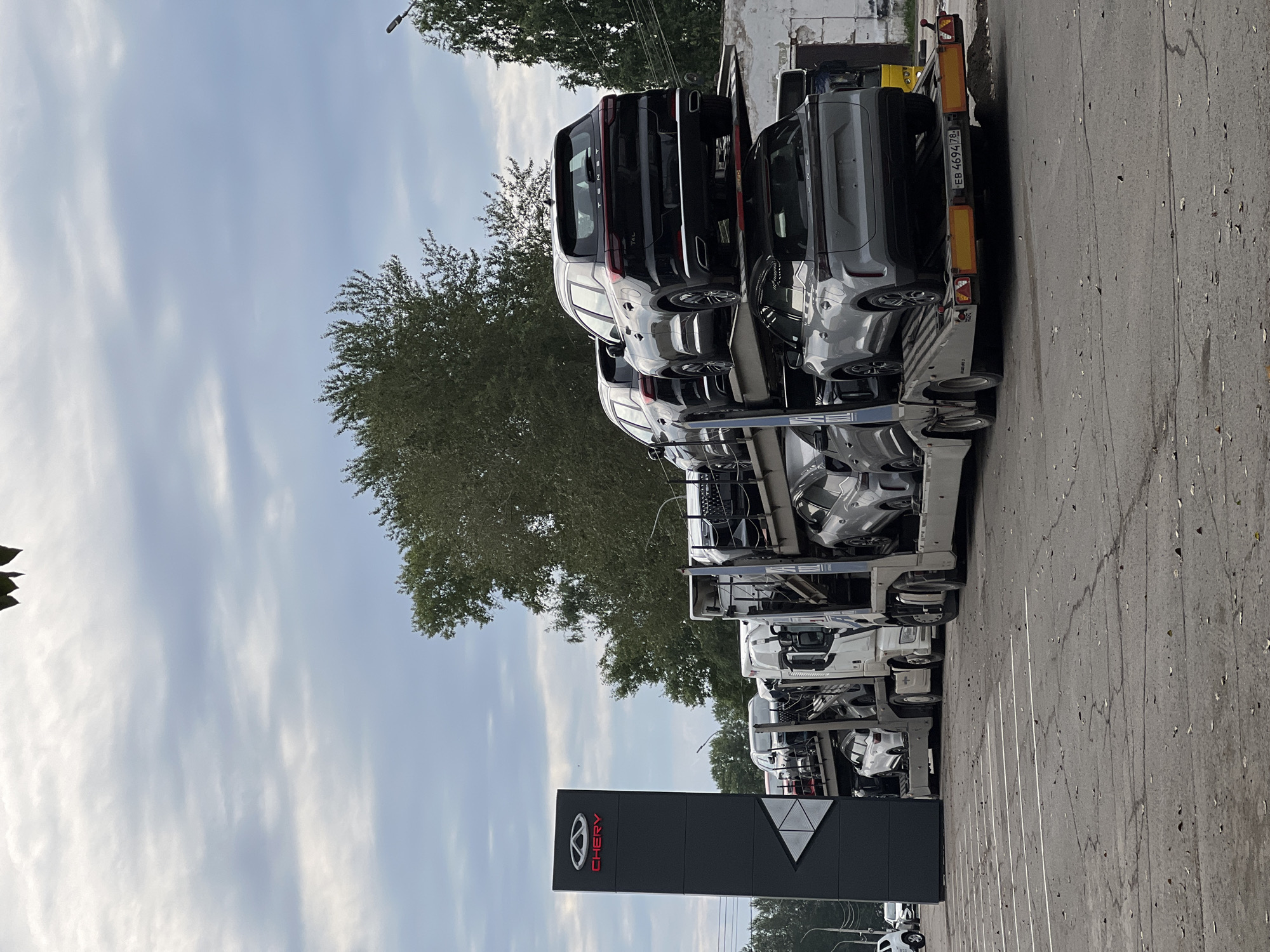 A car carrier truck is parked in a lot outside a Chery dealership in Izhevsk, Russia. The truck is loaded with several Chery vehicles on multiple levels. A tall, dark gray sign with a red Chery logo stands to the left of the truck. The sky is overcast with gray clouds. The ground is asphalt with white parking lines.