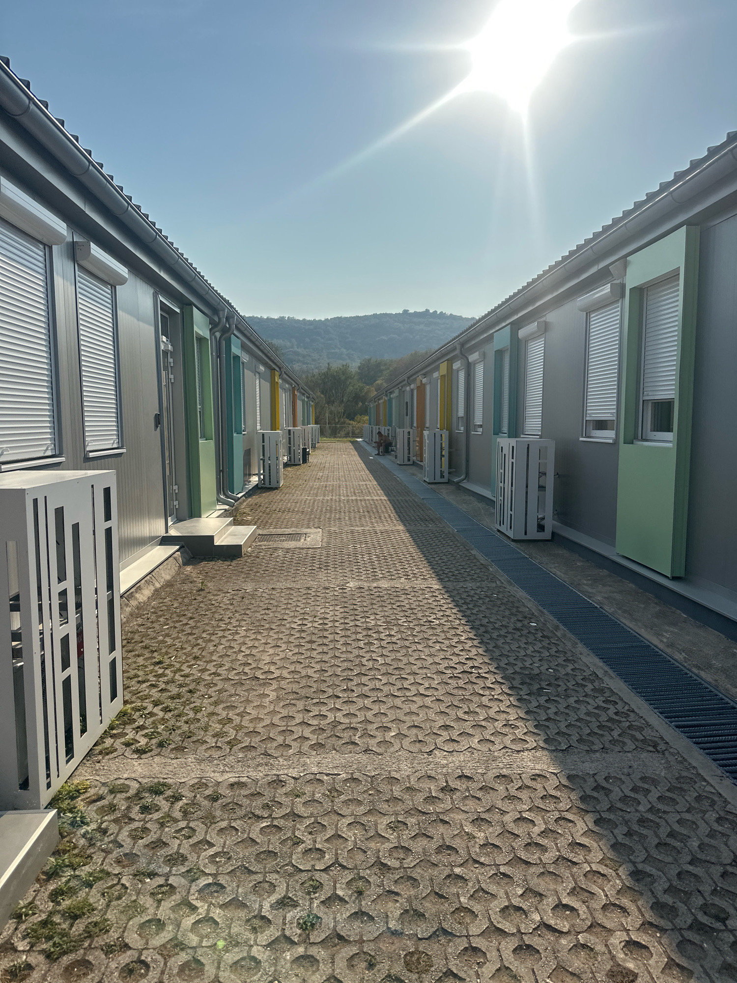 The image shows a perspective view down a paved walkway between two rows of prefabricated modular buildings in Radanovići, Montenegro. The buildings appear to be part of a new construction or development, with uniformly designed facades featuring grey paneling and brightly colored accents on the doors and window frames. Each unit has a white roller shutter over the window. Outdoor air conditioning units are visible in front of each building. The walkway is paved with interlocking concrete blocks, with some grass growing in the gaps. A metal grate runs along the edge of the walkway on the right side. The sky is clear and bright with the sun prominently visible, casting long shadows. In the distance, a gently sloping, tree-covered hill forms the backdrop. There are no visible people in the immediate foreground, but there are two figures seated near the buildings in the far distance on the right side. The overall scene suggests a well-organized, modern facility, possibly for residential or commercial purposes, under bright daylight conditions.