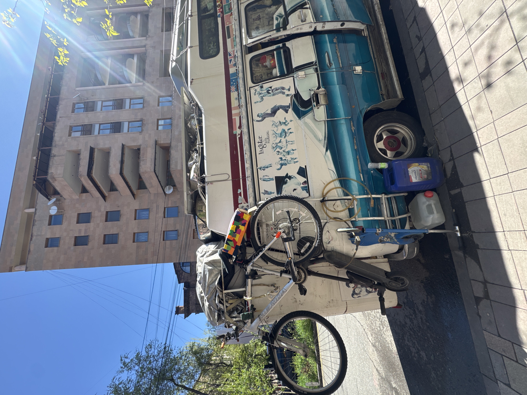 A brightly lit, vertical shot captures a distinctive, highly customized blue and white vehicle, likely an old double-decker bus or a large camper van, parked on a tiled sidewalk under a clear blue sky in Yerevan, Armenia. The vehicle is extensively decorated; a strip of international flags runs along its side, and a large graphic features various figures in action poses with the prominent text "Laughter is the best medicine."
Attached to the rear of the vehicle is a silver Merida bicycle, and below it, an electric scooter. A blue jerry can, possibly for fuel or water, with some unreadable text and a clear plastic jerry can sit near the rear wheel on the ground. Various wires, hoses, and ropes are visible around the vehicle's exterior. A gray tarp covers equipment on what appears to be a roof rack or rear platform.
In the background, a tall, multi-story building with a light-colored, possibly concrete or stone, facade dominates the upper portion of the frame. It features numerous windows and distinctive, large ventilation or cooling units on its higher floors. Strong sunlight from the upper left creates lens flare and casts sharp shadows across the sidewalk, indicating a bright, sunny day. Green foliage from nearby trees is visible towards the bottom left. The overall scene suggests a nomadic or adventurous lifestyle within an urban setting.
