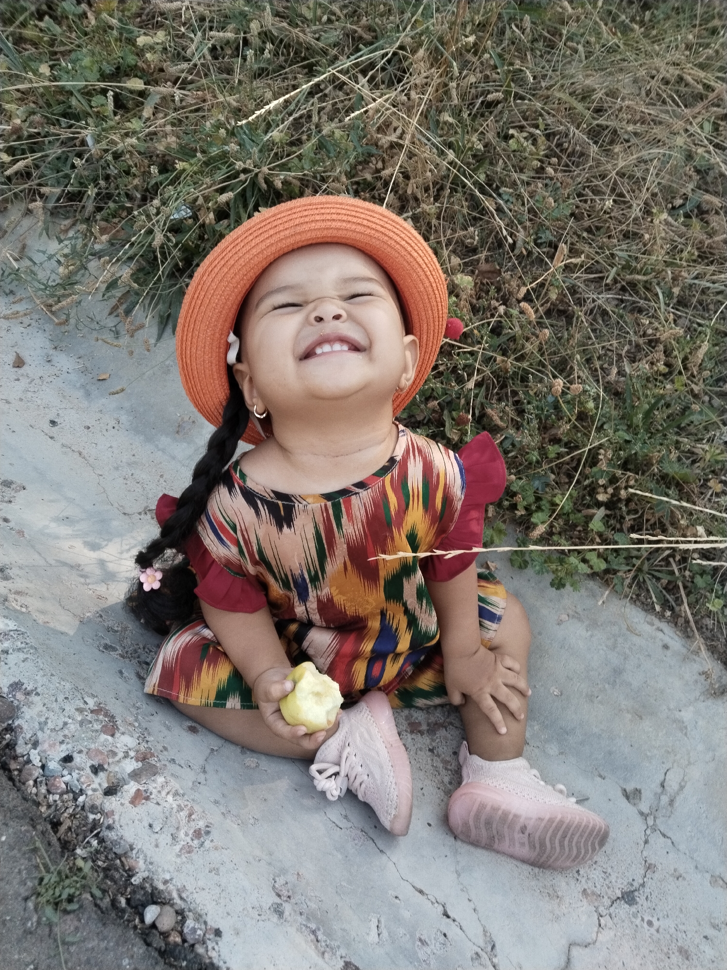 A young girl is sitting on a concrete surface in Akata, Uzbekistan, with a dry grassy area behind her. She is wearing an orange hat, a colorful patterned dress with red sleeves, and pink sneakers. The girl is holding a piece of light-colored fruit in her right hand and looking upwards with a wide, happy smile. Her dark hair is braided with a pink flower. The concrete surface shows signs of wear and tear, with cracks and scattered small stones. The environment suggests an outdoor setting, possibly a park or a less-maintained area.