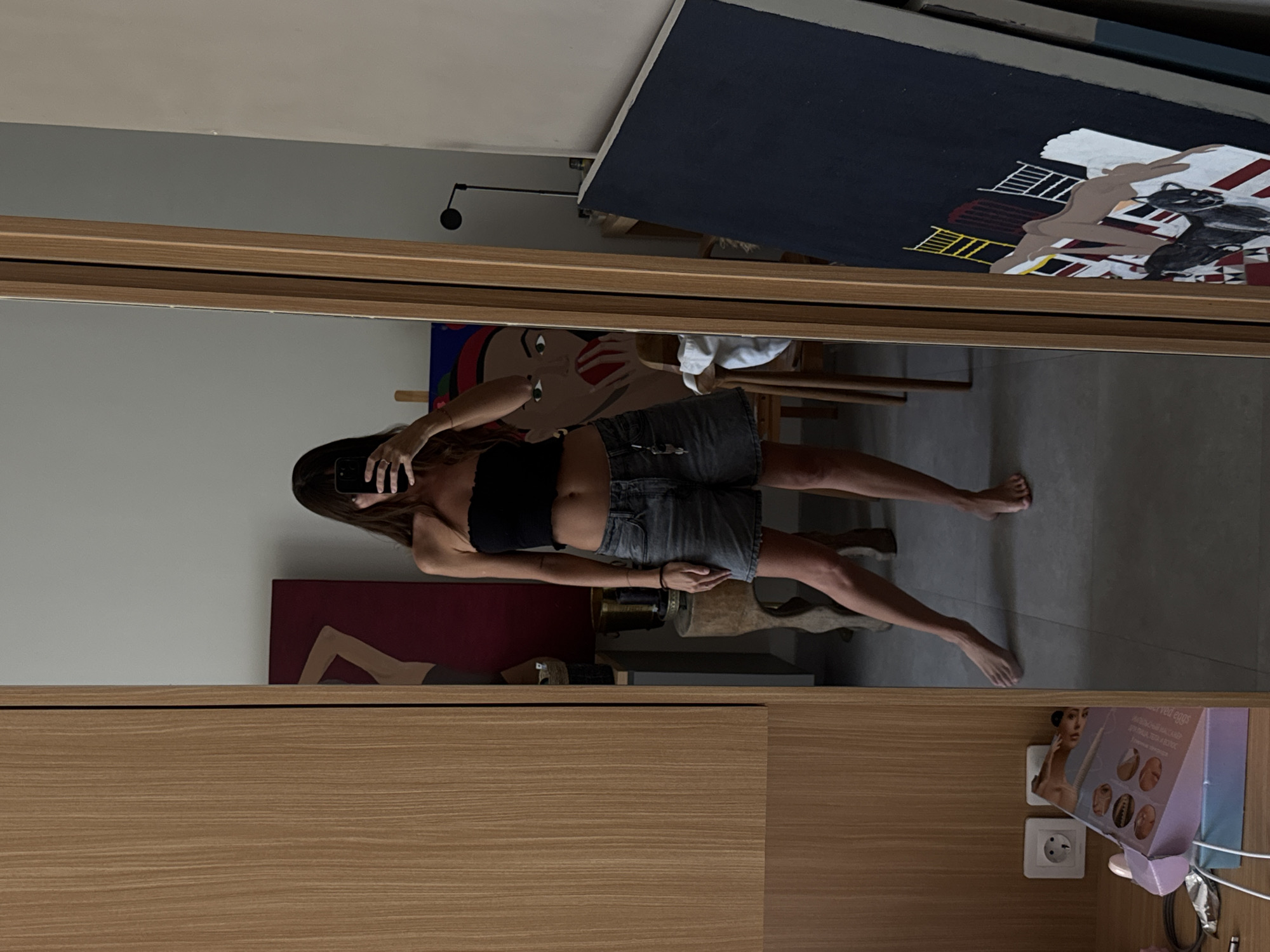The image shows a woman taking a mirror selfie inside what appears to be a residential space. The mirror is framed with light-colored wood, possibly part of a closet or cabinet. The woman is wearing a black tube top and gray denim shorts. She is holding a smartphone up to take the photo.
Behind the woman in the mirror's reflection, there are various objects including paintings, one with a cartoon-like face and another with figures, and what seems to be art supplies or furniture. Near the bottom of the frame, a box of cosmetic products is visible along with an electrical outlet. The location is indicated to be in North Kuta, Indonesia, although this is not directly verifiable from the image alone.