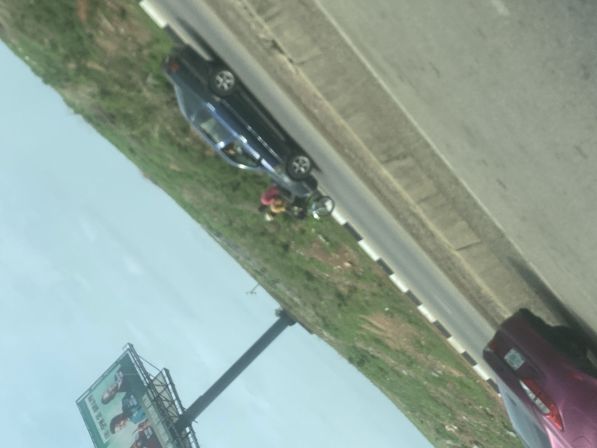 The image depicts an outdoor scene along a roadside or highway in Gosa, Nigeria, under what appears to be daytime conditions with a light blue, cloudy sky. The photograph is oriented with a significant counter-clockwise rotation.
In the mid-ground, a dark blue sedan is stationary or parked on a grassy verge next to a paved road. Near the driver's side of this blue car, at least one person wearing a red top and yellow lower garment is visible, standing next to a motorcycle. The exact nature of their activity is unclear, but they appear to be gathered around the car and motorcycle. The road itself is multi-laned, with clear asphalt surfacing and a white dashed line marking the shoulder or lane division.
To the far right, the rear section of a magenta or purple car is partially visible, suggesting the photo was taken from another vehicle. On the left side of the frame, a large billboard is prominently displayed, mounted on a sturdy black pole. The billboard features images of people and includes text, though the specific words are unreadable due to blurriness and resolution. The landscape surrounding the road is characterized by green grass and some scattered shrubs. No distinct time of day other than daytime can be determined, and the weather appears clear.