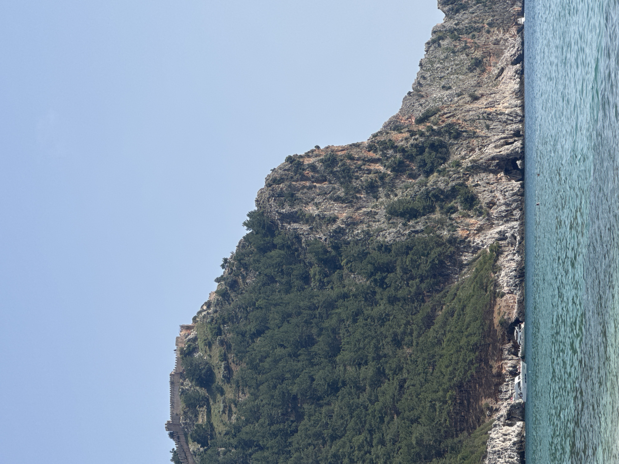 The image depicts a coastal scene in Alanya, Turkey. The frame is divided between a clear, pale blue sky on the left and the turquoise sea on the right. A rocky, mountainous coastline occupies the central area of the image, featuring vegetation, varying from sparse growth at the summit to dense, dark green trees at the base. Along the side of the mountain, a stone structure resembling an old wall or fortification is visible, suggesting a historical or architectural element on the landscape. Moored at the base of the mountain are several white boats. The overall impression is a scenic view combining natural and built environments.