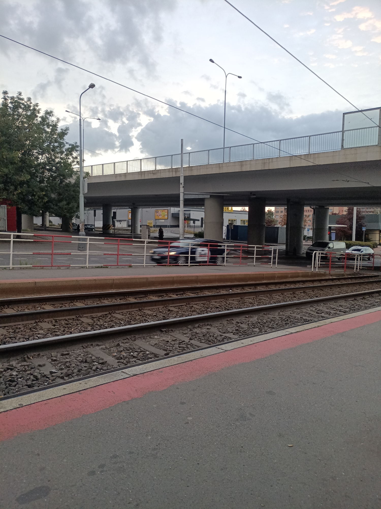 The image shows an outdoor scene with a street, railway tracks, and an overpass. A dark blue car is moving on the street from left to right, blurred due to motion. A black van and a silver car are parked on the right side of the street, under the overpass. A white fence with red and white stripes separates the street from the railway platform. The railway tracks are made of gravel and metal rails, with a red line painted on the adjacent platform. A few trees are visible on the left side of the image. The sky is cloudy and overcast. The scene appears to be a transportation hub in an urban environment. This location is likely to be Prague, Czechia, given the context of the surroundings.