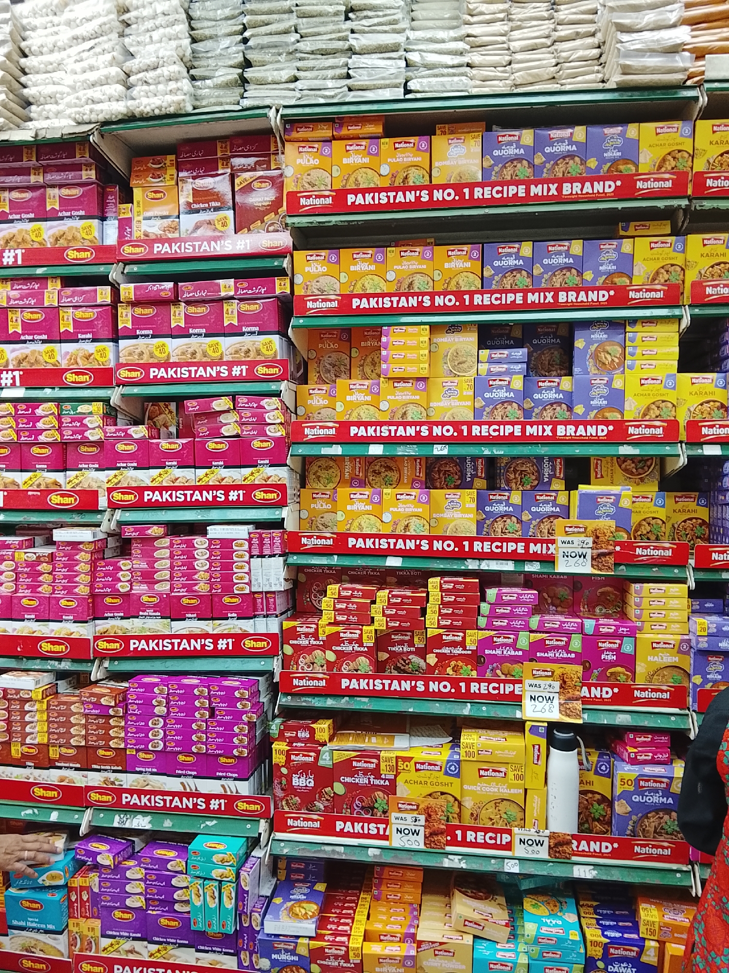 The image shows a grocery store aisle in Gulgasht Colony, Pakistan. Shelves are stocked with boxes of Shan and National brand recipe mixes. The top shelves hold large bags of unidentifiable food items. The Shan mixes are predominantly pink and brown boxes, while the National mixes are mostly yellow and blue. Signs reading "PAKISTAN'S #1" are placed on the Shan products. Promotional discounts are displayed on many of the boxes. A white water bottle is visible on one shelf. A hand reaches for a package on the bottom shelf. A person wearing a red patterned shirt is partially visible on the right side of the image.