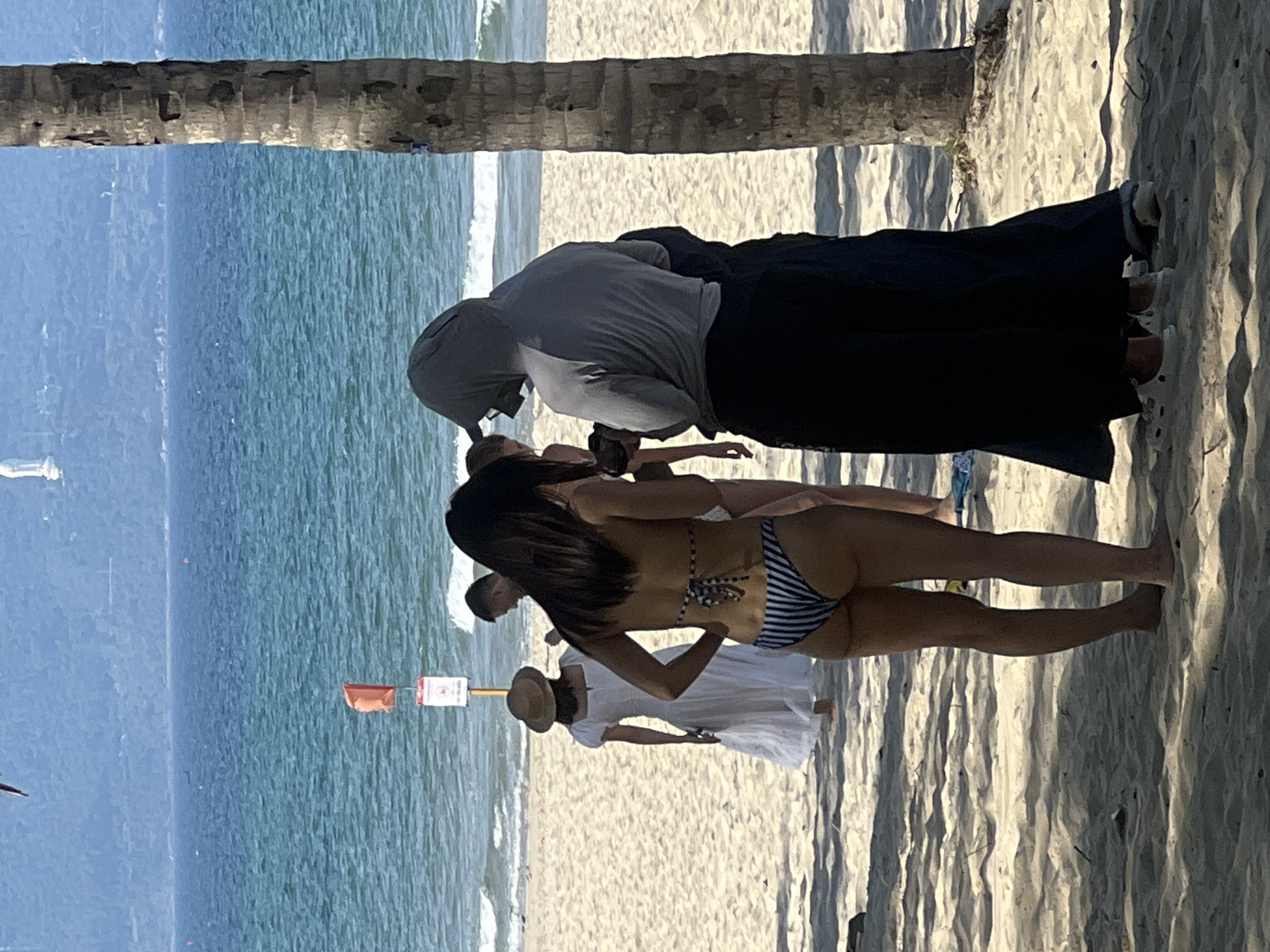 This image captures a sunny day at a beach, likely in Đà Nẵng, Vietnam, given the presence of a palm tree and the general coastal landscape. In the foreground, a person wearing a striped bikini stands facing away from the camera, with their hands on their hips. To their left, another individual is partially visible, also facing away and dressed in dark clothing, including a hooded top and dark trousers. Further in the background, a woman in a white dress and a straw hat is walking away from the camera on the sandy beach. Behind her, a man is also visible. The turquoise ocean stretches out to the horizon, with small waves lapping the shore. A red flag and a signpost are situated in the shallow water near the beach. The scene is illuminated by bright sunlight, casting shadows on the sand.