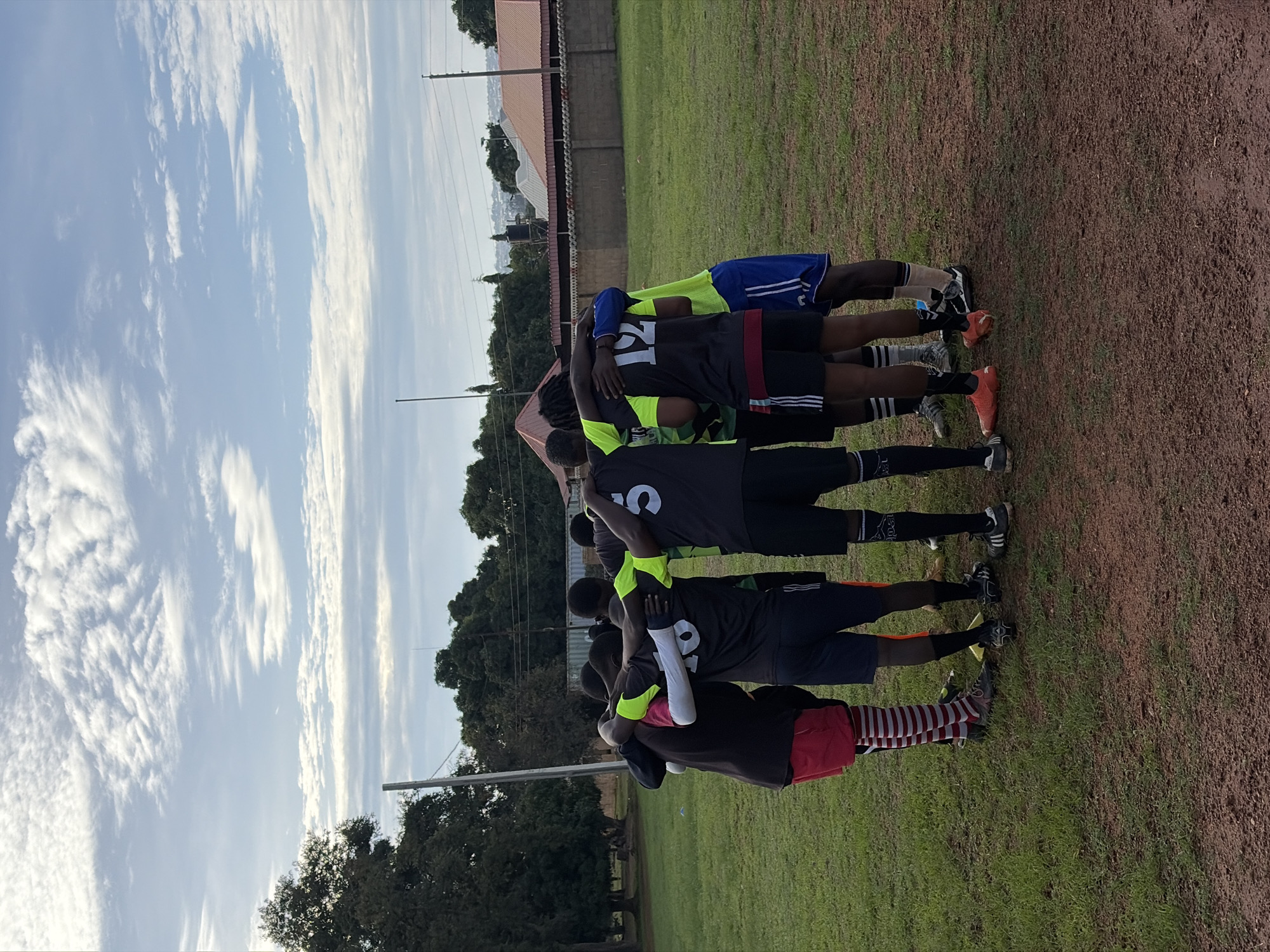 A group of eight young men, appearing to be a sports team, are captured from behind in a close huddle on a partially grassed, reddish-brown dirt field, located in Jos, Nigeria. Their arms are wrapped around each other's shoulders and backs, indicating teamwork or a strategic discussion. The individuals are wearing various soccer kits: black shorts, dark jerseys with bright neon yellow accents, and athletic footwear. Visible jersey numbers include "18" or "1B", "12", and "5". One player on the far left wears distinctive red and white striped socks and red shorts. In the midground, dense green trees frame the left side of the scene. On the right, a low, light-colored concrete or brick wall, topped with spherical decorative elements, separates the field from distant residential structures with red roofs. A black water tank on a stand is visible above the wall. Two tall, slender utility poles rise into the sky on either side of the field. The sky above is light blue with scattered white, feathery clouds.
