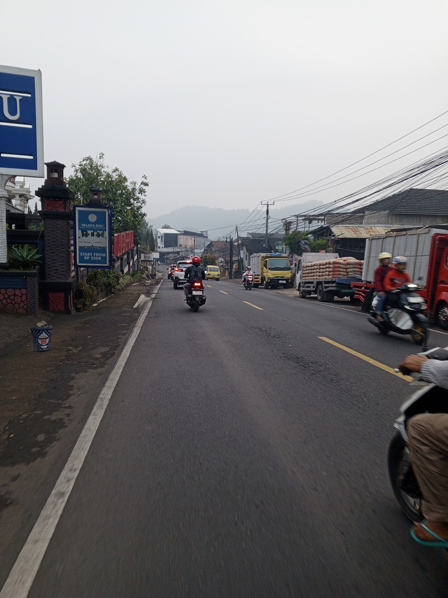 A busy street scene in Dambulla, Sri Lanka, features a variety of vehicles and activity. In the foreground, a motorcycle is partially visible, its rider wearing brown pants and turquoise flip-flops. The road is asphalt, marked with yellow and white lines. Several other motorcycles, cars, and trucks are also on the road, with some trucks appearing to be carrying sandbags. Buildings and power lines line the street, and a hilly landscape is visible in the distance under an overcast sky. A blue sign with white lettering and a blue trash can are present on the left side of the road, near a decorative gate and vegetation.