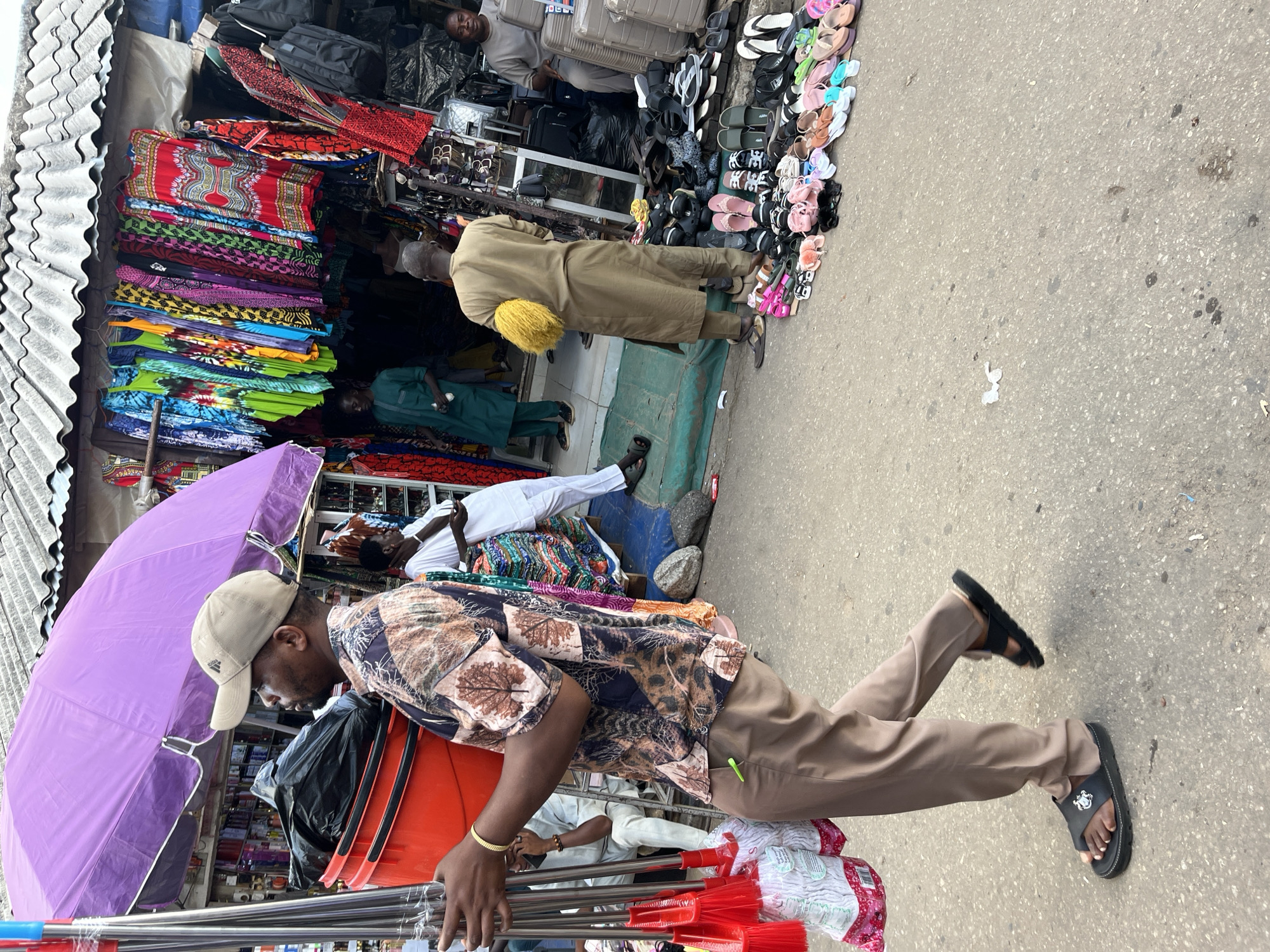 In Abuja, Nigeria, a busy marketplace unfolds. The scene is dominated by a stall overflowing with vibrantly colored textiles, showcasing intricate patterns in red, blue, green, yellow, and purple. Adjacent to the fabrics, a display of assorted flip-flops and sandals in various colors and styles is neatly arranged along the ground.
Several individuals are present. In the foreground, a man wearing a patterned shirt, beige pants, and a beige baseball cap is carrying a bundle of red-bristled cleaning tools, possibly brooms or mops. He is walking towards the viewer. Another man, dressed in a long, light brown garment, stands near the textile stall, holding a yellow fluffy object. Further in the background, a person in a green traditional outfit is seated. Another man in a white garment is also visible.
The setting appears to be an outdoor market, with a corrugated metal roof structure on the left. The ground is paved with asphalt, showing some signs of wear and tear. The lighting suggests it is daytime, possibly overcast due to the soft, diffused light. The overall atmosphere is one of commerce and daily life in a Nigerian urban environment. The subjects seem engaged in their activities, with no overtly discernible emotions. No visible text is present in the image.