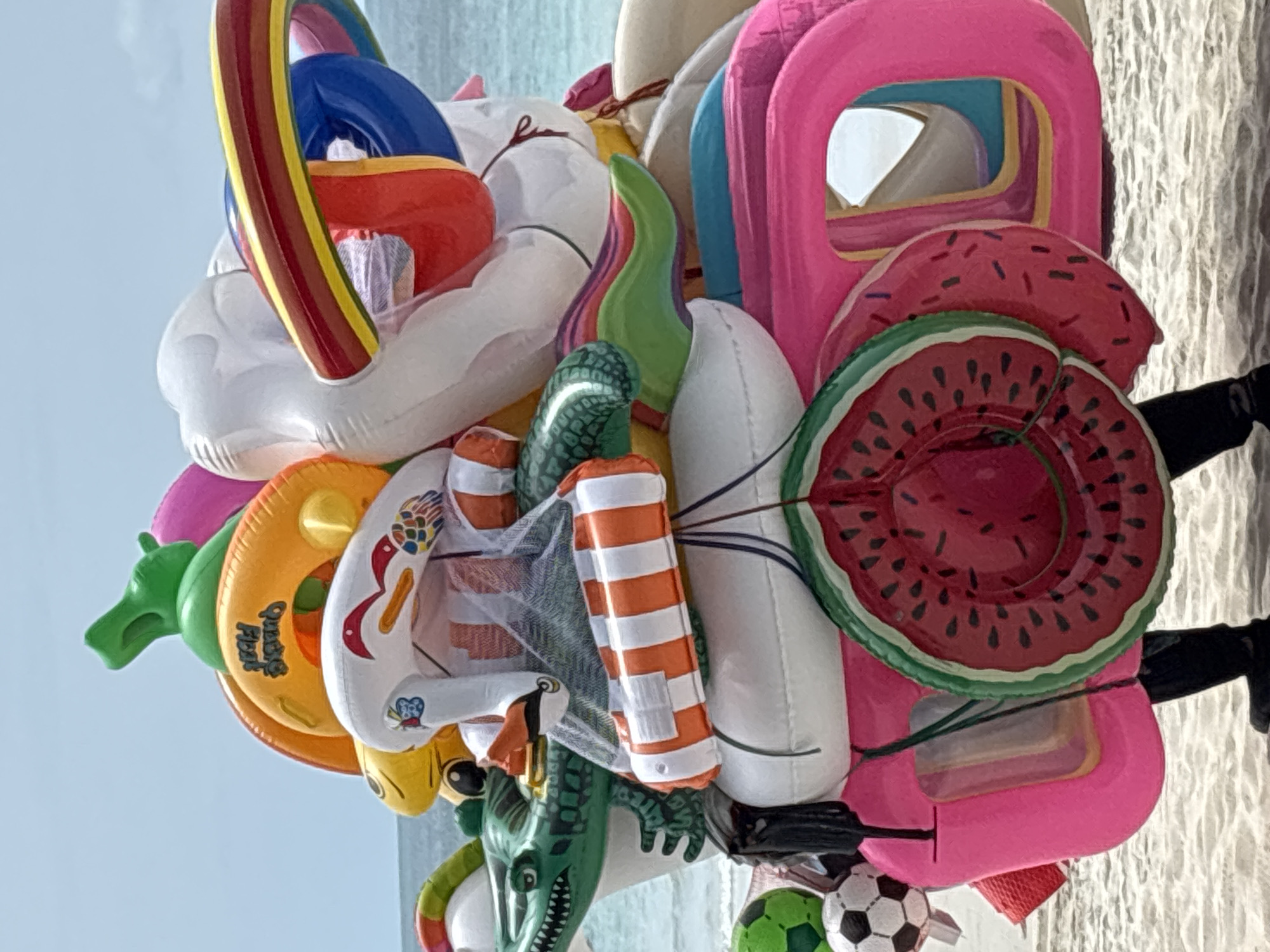 A person wearing black pants and black shoes stands on a sandy beach with inflatable water toys. The person is carrying a large assortment of colorful inflatable toys, including a green alligator, orange rings, a white swan with red accents, a striped orange and white inflatable, watermelon-shaped floats, a pink inflatable, and a soccer ball. In the background, the ocean and a clear sky are visible. The scene is likely in Baan Chaweng, Thailand, a popular beach destination known for its vibrant atmosphere and tourist activities.