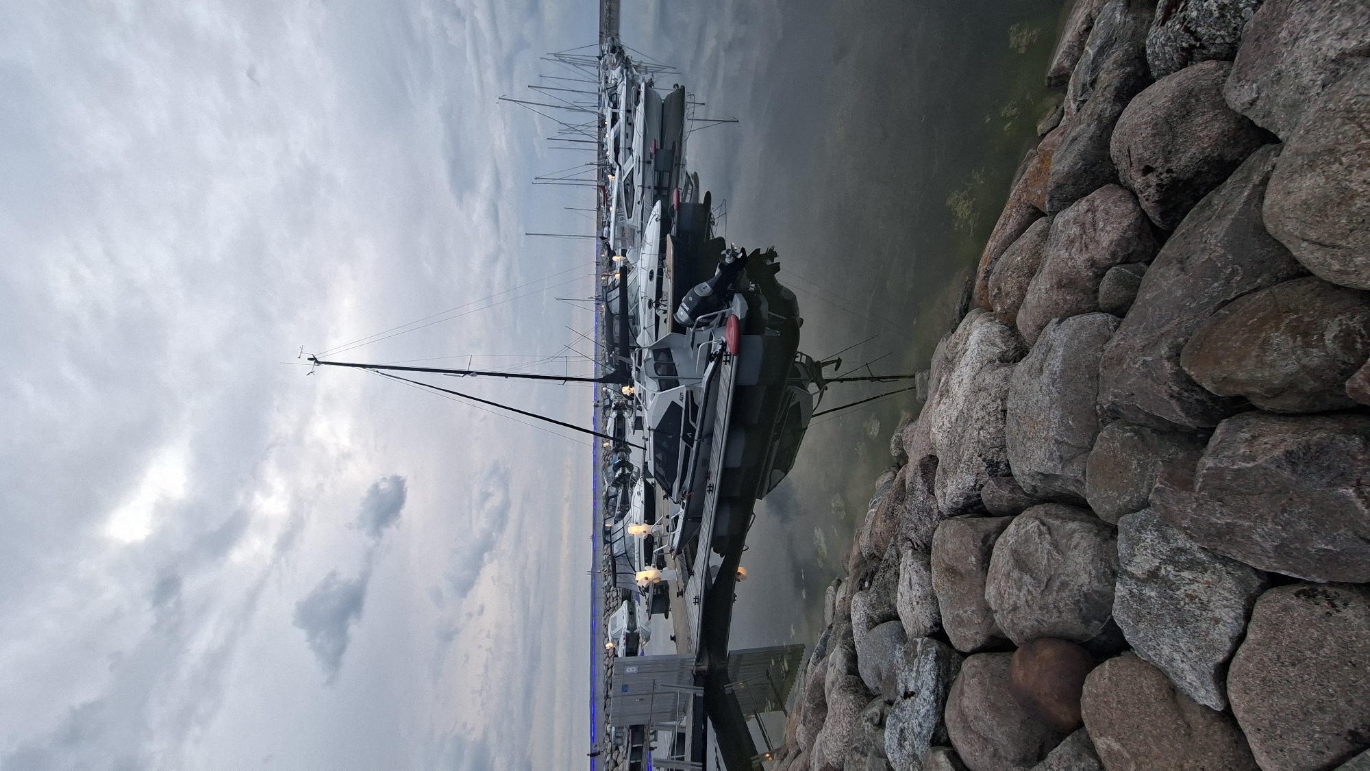 This media file depicts a marina in Tallinn, Estonia, likely during the early evening or late afternoon given the dim lighting and cloudy sky. The scene is dominated by a row of yachts and boats docked along a wooden pier. In the foreground, a large stone embankment lines the edge of the water, providing a textured and natural contrast to the man-made vessels.
The sky is overcast with diffused light, suggesting a cloudy or potentially drizzly day. The water is calm, reflecting the boats and the muted sky. The boats themselves appear to be of various sizes, with some larger yachts showcasing sleek designs and masts. Lights on the pier and some of the boats are illuminated, adding a touch of warmth to the cool-toned environment. There are no people visible in the frame.
The overall mood is one of quiet repose, typical of a marina at the end of the day. The location cues, such as the style of the boats and the stone embankment, are consistent with coastal areas in Northern Europe. The image focuses on the interplay of the natural (stones, sky, water) and the man-made (boats, pier), creating a serene and atmospheric composition. There is no visible text in the image.