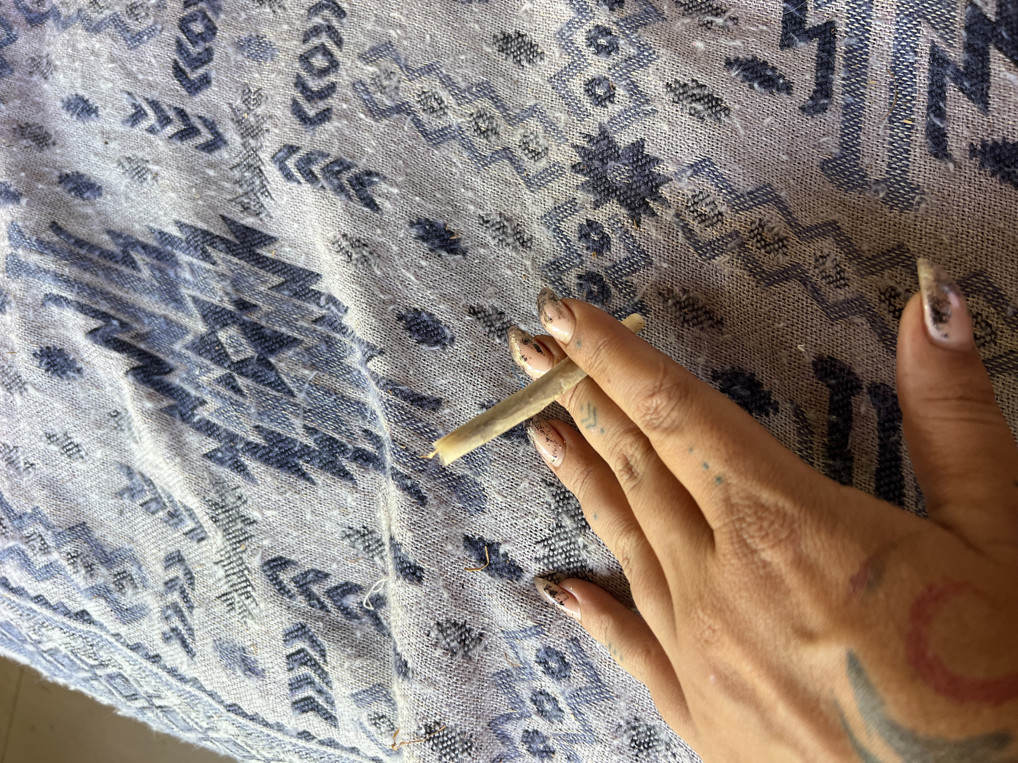 A hand holds a lit cigarette or hand-rolled joint over a patterned cloth. The hand has long nails with painted designs and a tattoo on the wrist. The cloth features a blue and gray geometric pattern. The location is not discernible from the image. The city of Sukawati, Indonesia, cannot be determined from the image.