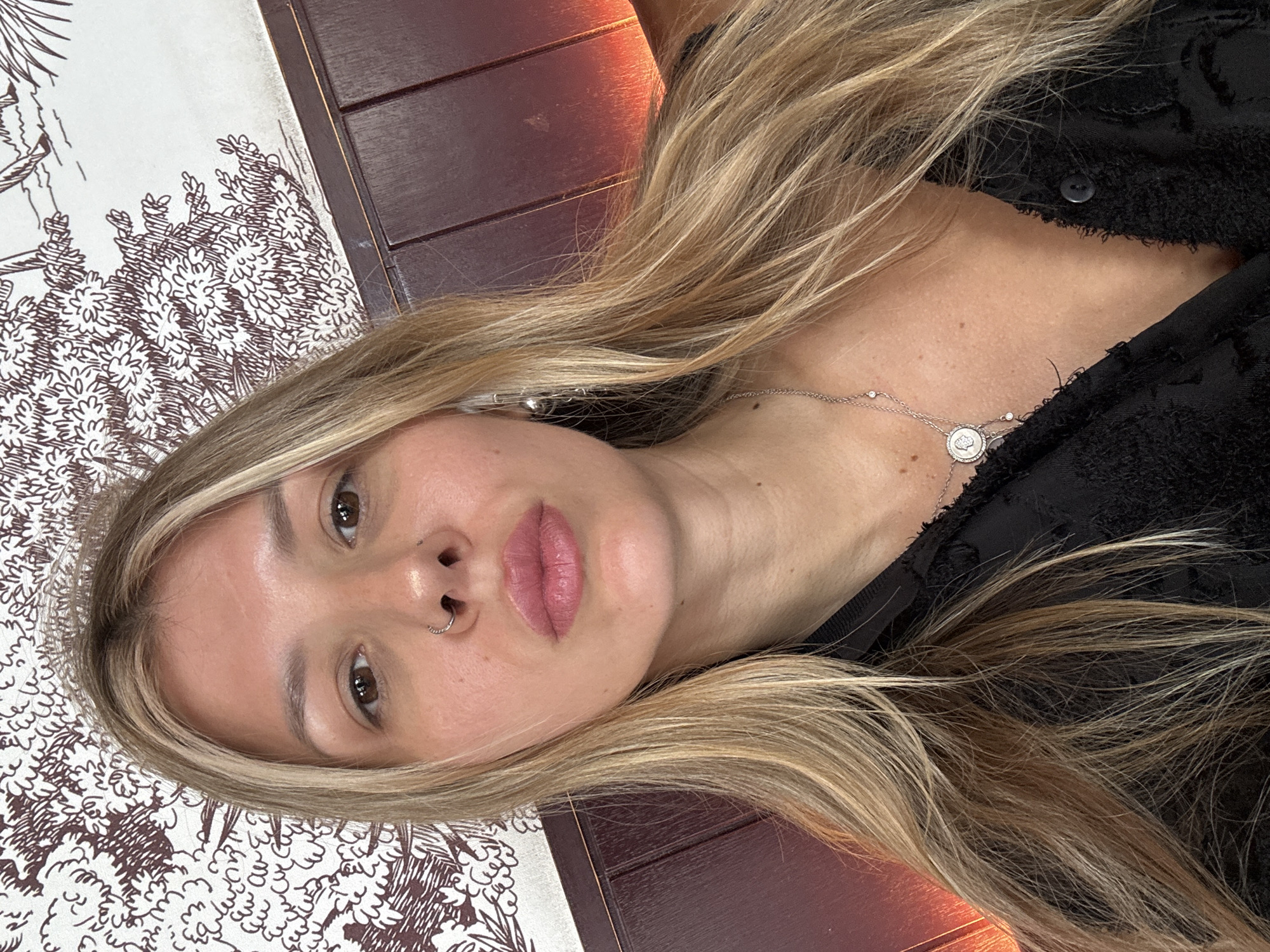 A medium shot shows a woman from the chest up, facing forward. She has blonde hair with streaks of lighter blonde, parted in the middle and falling past her shoulders. She is wearing a black textured shirt with a button closure. A delicate silver necklace with a circular pendant and small charms hangs around her neck. She has a nose ring and is wearing small, silver hoop earrings. Her makeup is subtle, with emphasis on her lips, which are painted a rosy pink. The background consists of patterned wallpaper with brown line drawings of foliage and what appears to be a wooden paneled wall with a warm, orange light reflecting on it.