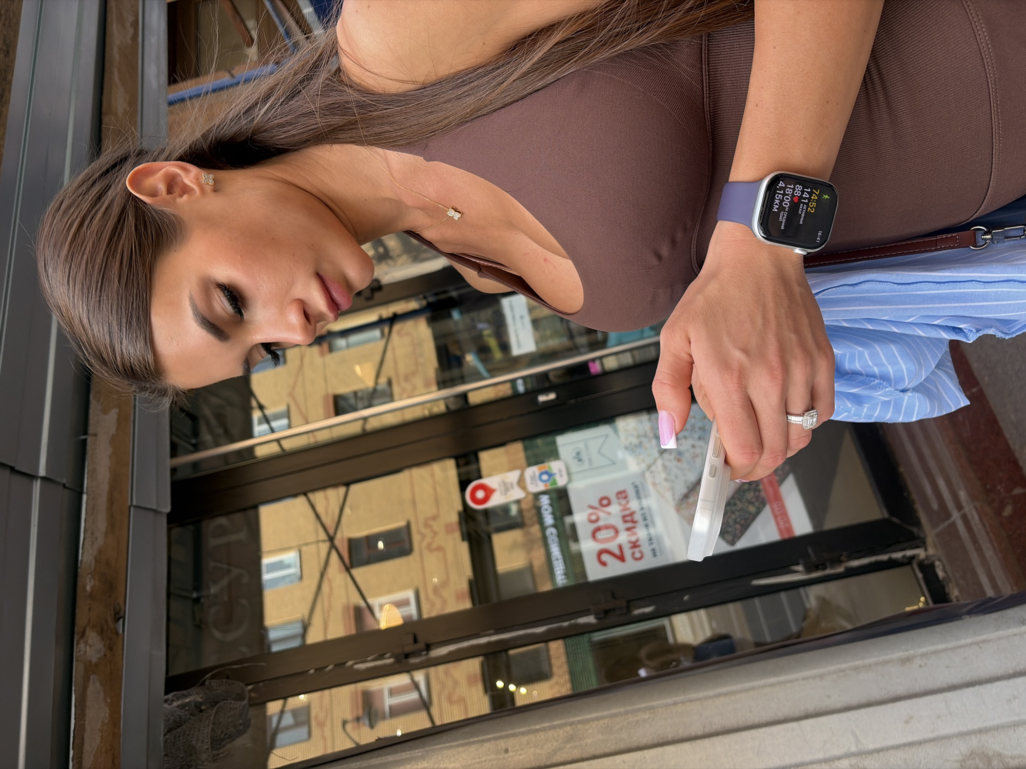 A young woman with long, brown hair pulled back in a ponytail is depicted in a medium shot, standing outdoors in the city of Saint Petersburg, Russia. She wears a form-fitting brown athletic top and a delicate gold necklace with a butterfly-shaped pendant. Small, gold butterfly stud earrings are visible in her ears. Her left wrist displays an Apple Watch with a purple band, showing Cyrillic text and workout data: a timer of 74:52, 141 kcal, a heart rate of 88 bpm, an average pace of 18:00, and a distance of 4.15 km, with the current time as 16:41. She holds a white smartphone, encased in a clear cover, in both hands, looking down at its screen. Her right hand sports a prominent diamond ring on her ring finger, and her fingernails are manicured with a white tip (French manicure). A light blue and white striped garment is draped over her left arm, with a portion of a brown bag strap also visible.
The woman stands next to a building with large, reflective glass doors and windows. On the glass, a red sticker shaped like a map pin is visible, along with another white sticker displaying "WOW" and an adjacent square icon. Below these, a clear red sign states "20% СКИДКА" (20% DISCOUNT) in Cyrillic. The glass reflects elements of the surrounding urban environment: a multi-story building with a light yellow/ochre facade and numerous windows, as well as a partial, reversed Cyrillic reflection that appears to be "СУР". To the left of the woman, a fluted architectural column is partially visible. The ground beneath her is a reddish-brown tiled or paved surface. Above the glass entrance, a metal awning structure and a raw wooden beam are present.