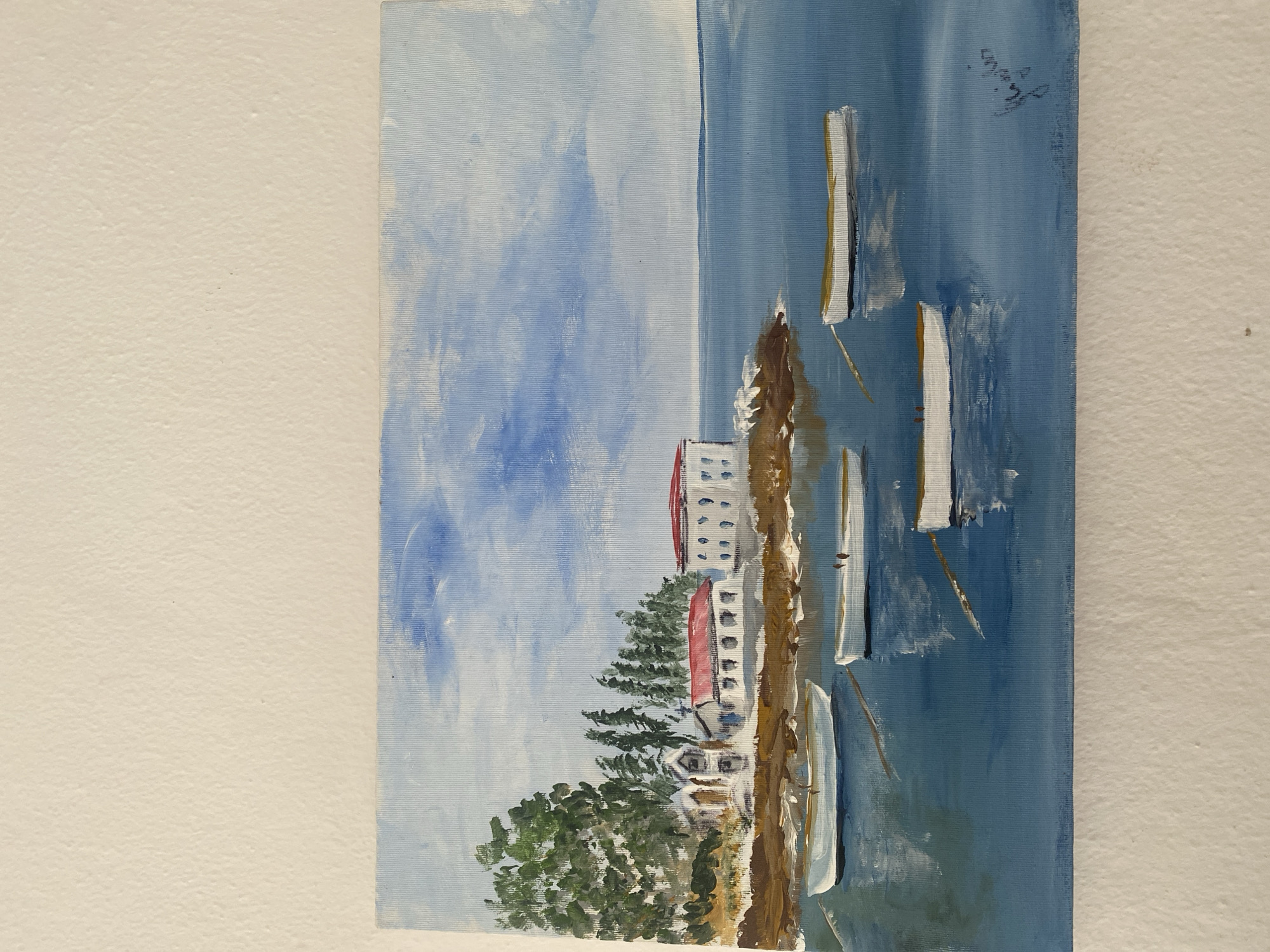 The image is a painting depicting a coastal scene with a building complex featuring red-roofed structures and smaller white buildings nestled among trees, adjacent to a shoreline. Several sailboats are portrayed on a blue body of water, with visible masts and white sails. A blue sky with white cloud formations is present above the landscape. A signature is located in the upper right corner. The location of the painting in Çekmeköy, Turkey is not depicted in the picture.