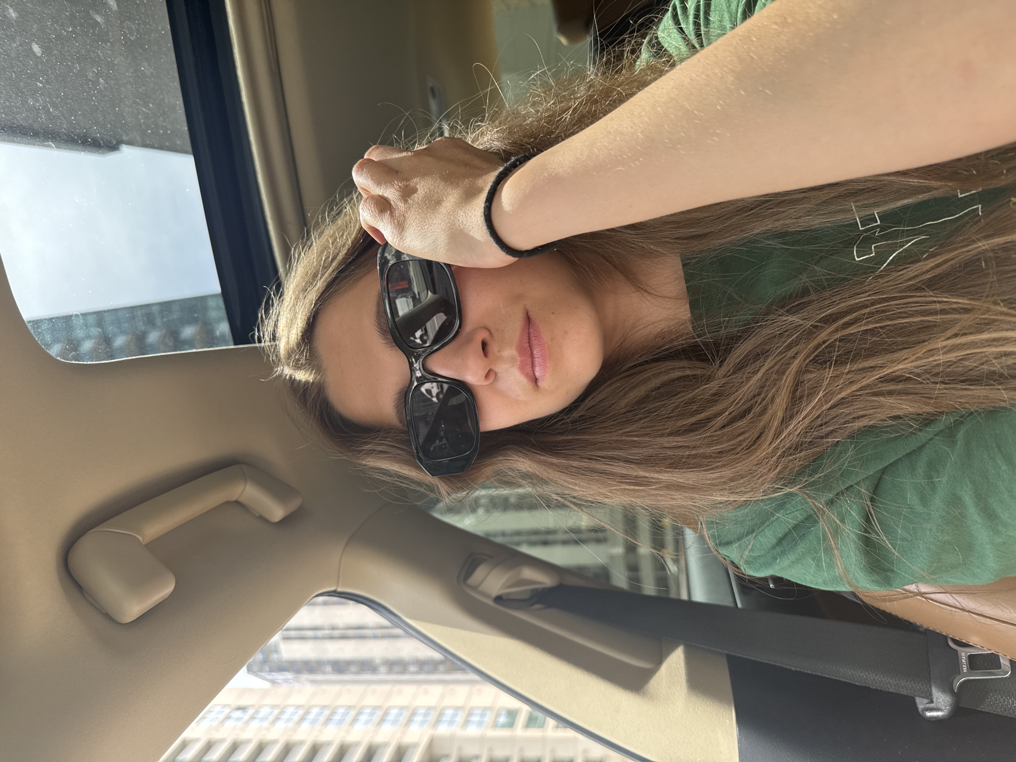 The image displays a young person, likely female, taking a selfie inside a car. The photograph is rotated 90 degrees clockwise, presenting the subject horizontally. She has long, light brown hair that falls over her shoulders and back. She is wearing a dark green t-shirt with a faint white graphic or logo visible on the left shoulder area, partially obscured by her hair. Black, rectangular sunglasses are pushed up onto her forehead, covering her eyebrows, while her lower face, including her nose and mouth, is visible. Her lips are slightly parted, and her expression appears neutral. Her right arm is raised, with her hand resting on her forehead, and a black elastic or bracelet is around her wrist.
The setting is the interior of a car, visible through beige headliner and trim on the upper section, including a grab handle on the left side. A black seatbelt with a silver buckle, featuring the partially visible text "HOVTM", crosses the lower right portion of the frame. Bright sunlight illuminates the scene, suggesting daytime. Through the car window on the left, blurred tall city buildings are visible, indicating an urban environment. The car window itself shows some dust. The image captures a casual moment, possibly while traveling in Ottawa, Canada.