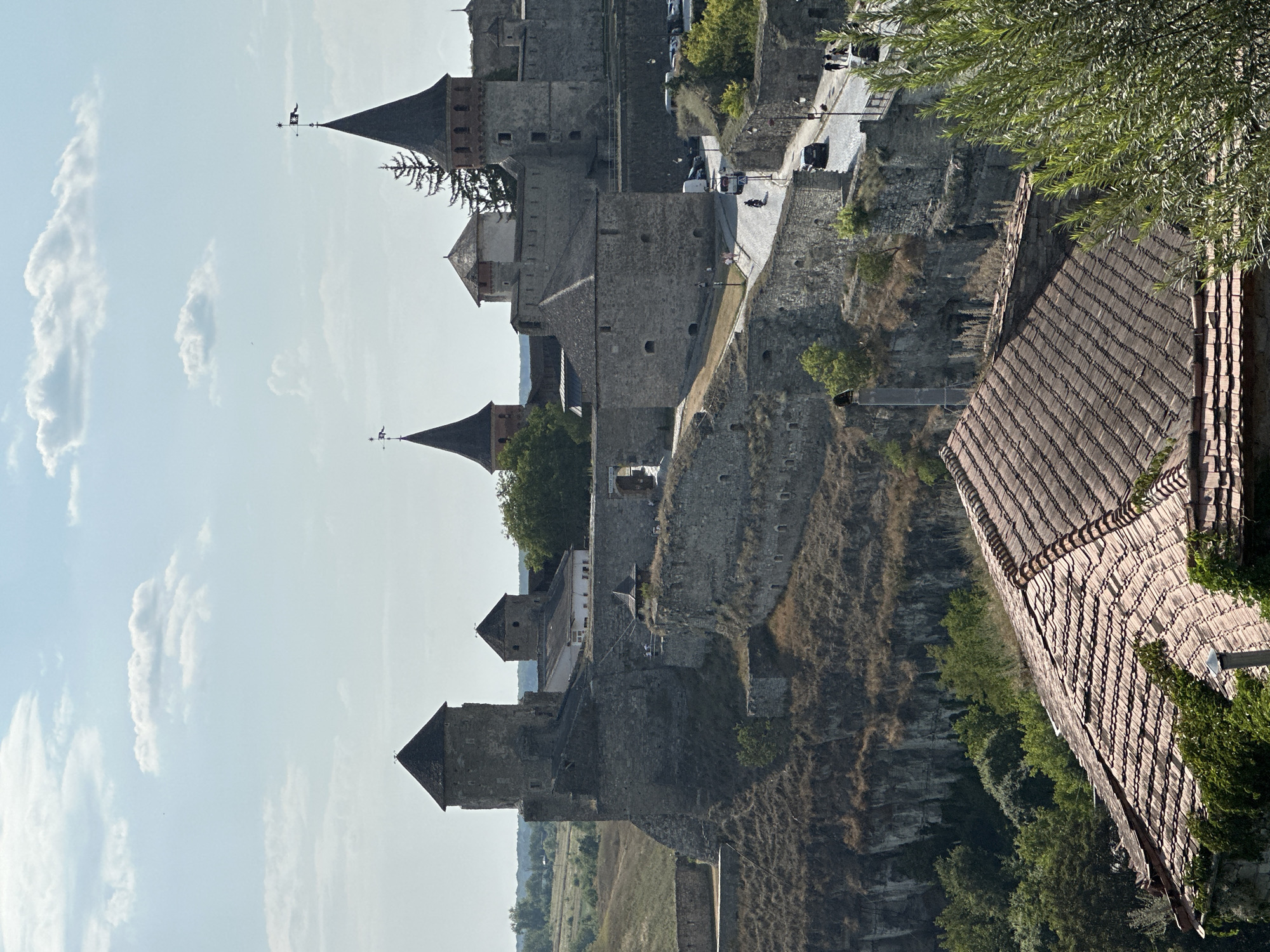 This is a daytime, outdoor, high-angle, full shot of the Kamianets-Podilskyi Castle in Kamianets-Podilskyi, Ukraine. The castle is a large, stone fortress with multiple towers and crenellated walls, perched on a rocky promontory. The terrain surrounding the castle is hilly and covered with dry grass and some scattered trees. In the foreground, a tiled roof of a building is visible, with green foliage growing on it. A paved road winds its way up to the castle, with several cars and a motorcycle on it. A few people are walking on the road. The sky is a light blue with scattered white clouds. The overall impression is one of a historic and imposing structure in a natural setting.