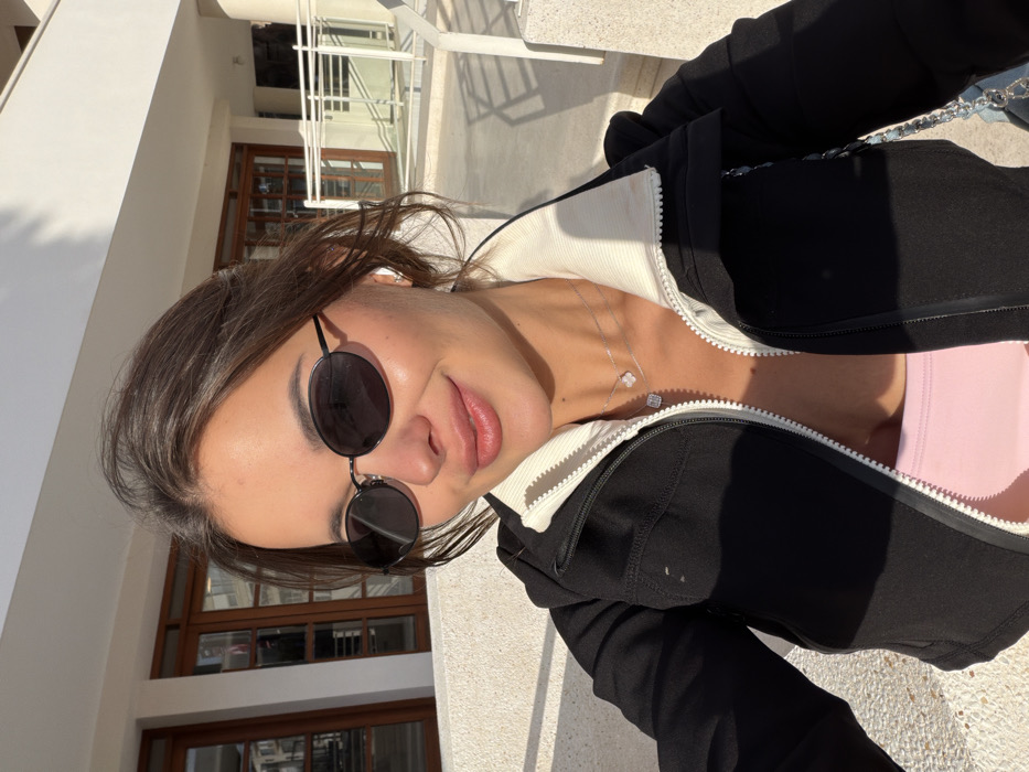 A single female subject is depicted in a sunlit outdoor close-up. She possesses dark hair and is wearing oval-framed dark sunglasses and a white wireless earbud in her right ear. Her lips are enhanced with a pink cosmetic. She is dressed in a black zip-up jacket featuring a white ribbed collar and visible white zipper, partially revealing a light pink garment underneath. Two silver necklaces are present: one with a four-leaf clover pendant and another with a square, possibly gem-set, pendant. A silver chain strap, likely from a bag, is visible on her left shoulder. The subject exhibits a slight smile and looks directly forward.
The background consists of a light-colored, possibly stone or concrete, parapet or ledge. Behind this, a modern white-walled building with large wooden-framed glass doors is visible. To the right, a white railing structure suggests a balcony or terrace. Strong overhead sunlight illuminates the scene, casting distinct shadows, indicative of daytime. The architectural style and environmental conditions are consistent with an urban setting in Tel-Aviv, Israel; however, a precise location within the city cannot be determined from the image.