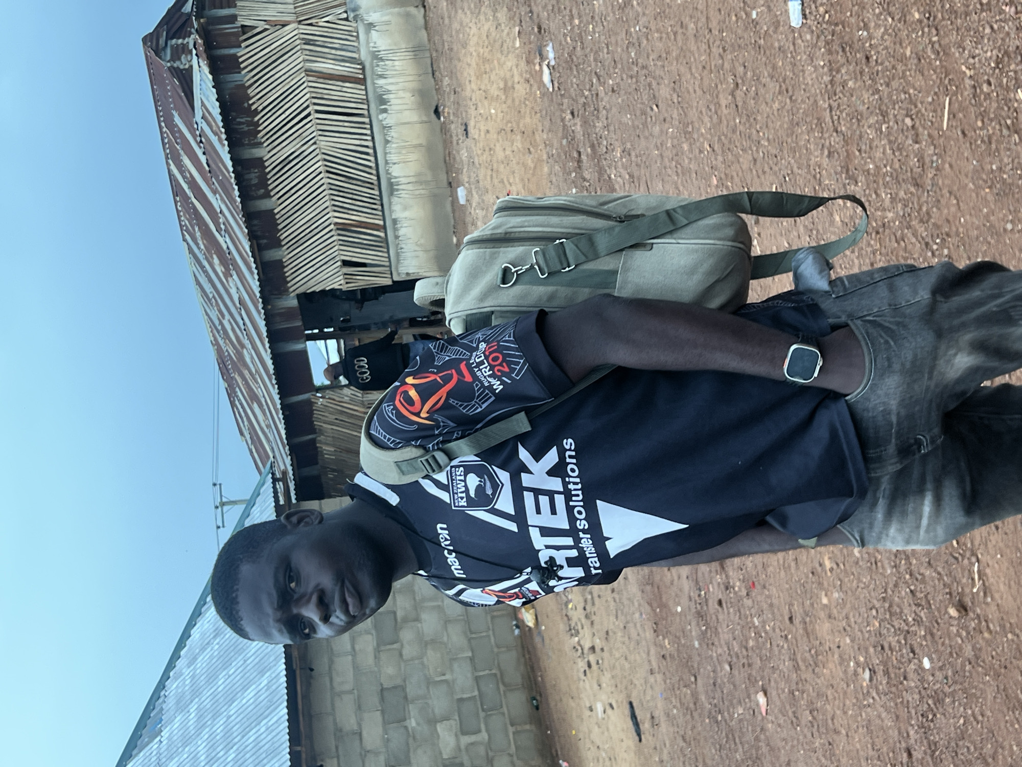 The image shows a young Black man wearing a black sports jersey with white and red accents, a green backpack, and gray jeans. He is standing in an outdoor environment with buildings in the background. The buildings appear to be constructed with corrugated metal roofs and brick or wood walls, suggesting a rural or semi-urban setting. The ground is covered in dirt and gravel. The man is looking towards the camera with a neutral expression. The sky is overcast. There is a person in the background wearing a jacket with the word "GOOD" on the back. The location is identified as Durimi, Nigeria.