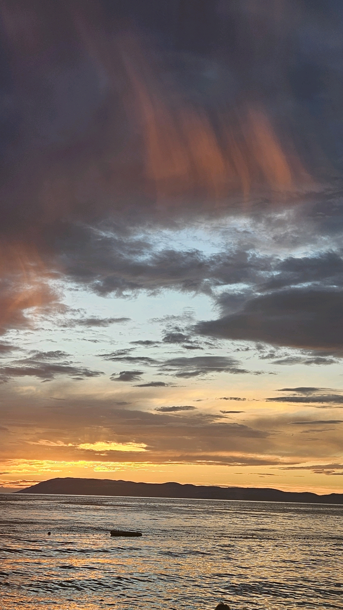 This is a vertical, eye-level shot of a sunset over the Adriatic Sea, as viewed from the coast of Općina Tučepi, Croatia. The sky is a dramatic display of color, with dark gray clouds dominating the upper portion, punctuated by streaks of orange and pink light, likely from the setting sun. Lower in the sky, lighter gray and blue clouds create a layered effect. The horizon is marked by the silhouette of a distant mountain range. The sea in the foreground is calm, with ripples reflecting the warm hues of the sunset, casting a golden sheen on the water. A small, dark object, possibly a buoy or a small boat, is visible in the water. The overall impression is one of a serene and beautiful natural landscape at dusk.