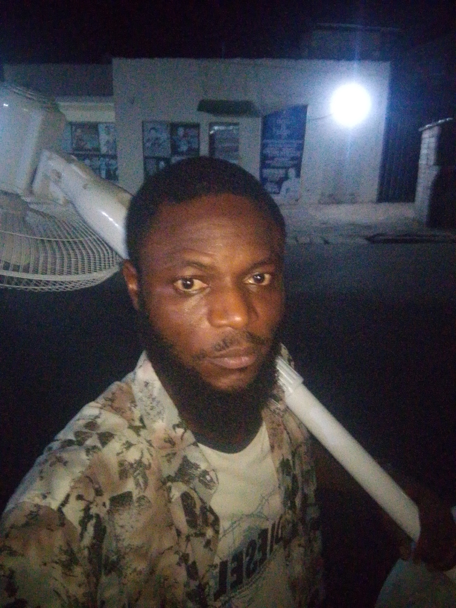A man with dark skin and a full beard stands in front of a building at night. He is carrying a disassembled white fan over his shoulder. The building has a bright light illuminating the front and windows. The location is the city of Unknown, Unknown.