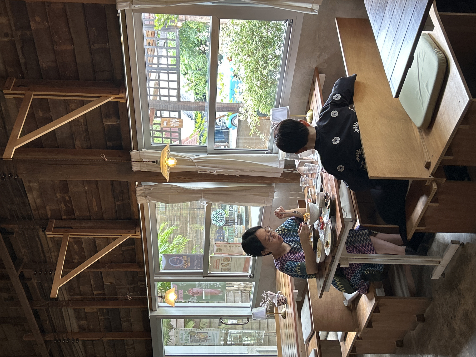 In Chiang Mai City Municipality, Thailand, two individuals are seated at a wooden table in a cafe. The person on the left, wearing glasses and a colorful patterned dress, is actively eating from a bowl with utensils. The person on the right, facing away from the camera and wearing a dark kimono-style garment with floral embroidery, is also seated at the table. Various dishes and bowls are spread across the table, suggesting a meal is in progress. The cafe's interior features exposed wooden beams and walls, with large windows offering a view of lush greenery and a blue vehicle outside. Natural light illuminates the space, complemented by hanging pendant lights. The seating arrangement consists of wooden benches with cushioned seats.