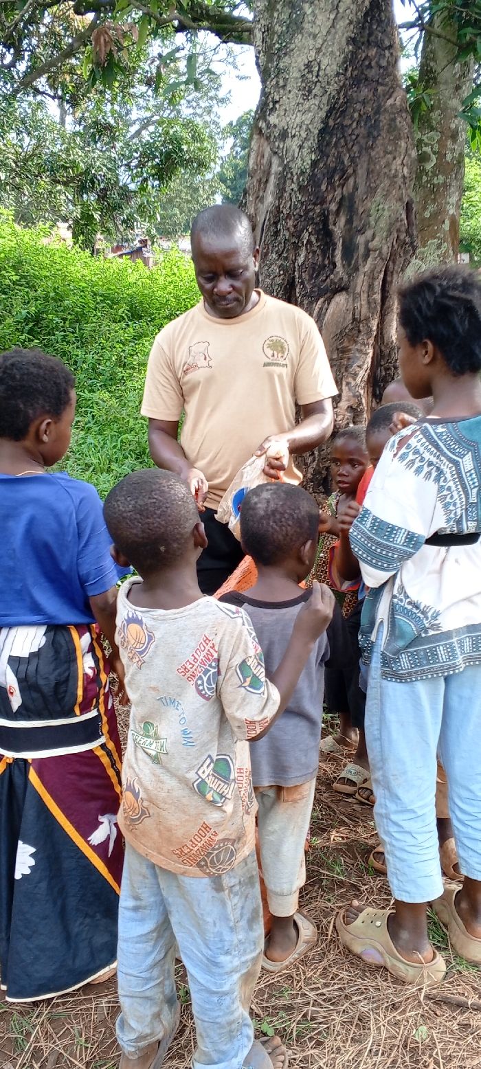 The image depicts an outdoor scene in the city of Ngalula, Democratic Republic of the Congo, featuring one adult male and a group of approximately seven children.
The adult male, of African descent with short, dark, receding hair, stands centrally, slightly hunched, directing his gaze downwards. He wears a tan short-sleeved t-shirt with two distinct logos on the front: a light brown, irregularly shaped graphic on the left chest and a circular logo featuring a tree design and the partial text "ARDECH..." on the right chest. He is engaged in an action involving a crumpled light-colored plastic bag held in his left hand, while his right hand reaches into or manipulates its contents, suggesting distribution or handling of items.
Clustered around the adult male are several children of varying young ages. From the left, a child with their back to the camera wears a blue t-shirt and a long skirt with multiple patterns, including black, maroon, yellow, white birds, and white flowers. Immediately to their right, another child, also seen from behind, wears a soiled light-colored t-shirt with various basketball-themed graphics and dirty light blue trousers, complemented by light-colored sandals. A third child, mostly obscured behind the second child, wears a dark t-shirt and light-colored trousers, with their left arm casually resting on the second child's back; this child also wears light-colored sandals. On the far right, a slightly older child faces the adult, wearing a white top with blue and black patterns and light blue trousers, along with light-colored sandals. Several other children are partially visible between the adult and the foreground children, with at least two additional faces visible, looking intently towards the adult male and his hands. One of these children wears a brightly patterned top.
The environment is an unpaved outdoor area. A large tree trunk with rough, textured bark dominates the right side of the frame. Dense green foliage and bushes form a backdrop behind the individuals, with hints of distant structures or additional trees visible beyond the immediate greenery. The ground is dry, reddish-brown earth with sparse dry grass or plant debris. Natural daylight illuminates the scene.