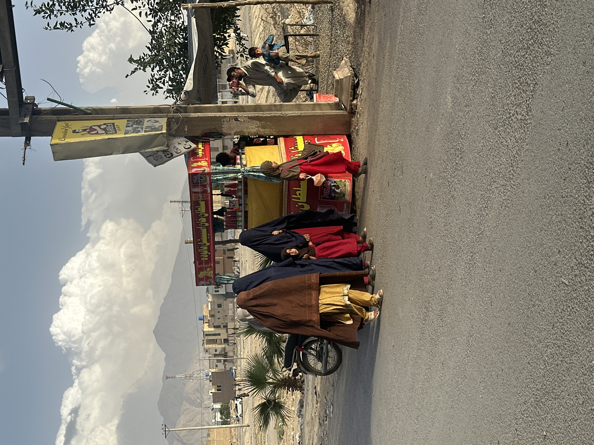 The image depicts a street scene in Quetta City Tehsil, Pakistan. Three women wearing burqas are seen on a motorcycle next to a beverage stand. Two men are standing near the stand, one drinking from a bottle or can. Mountains are visible in the background with buildings and a blue and cloudy sky. The street is made of asphalt.