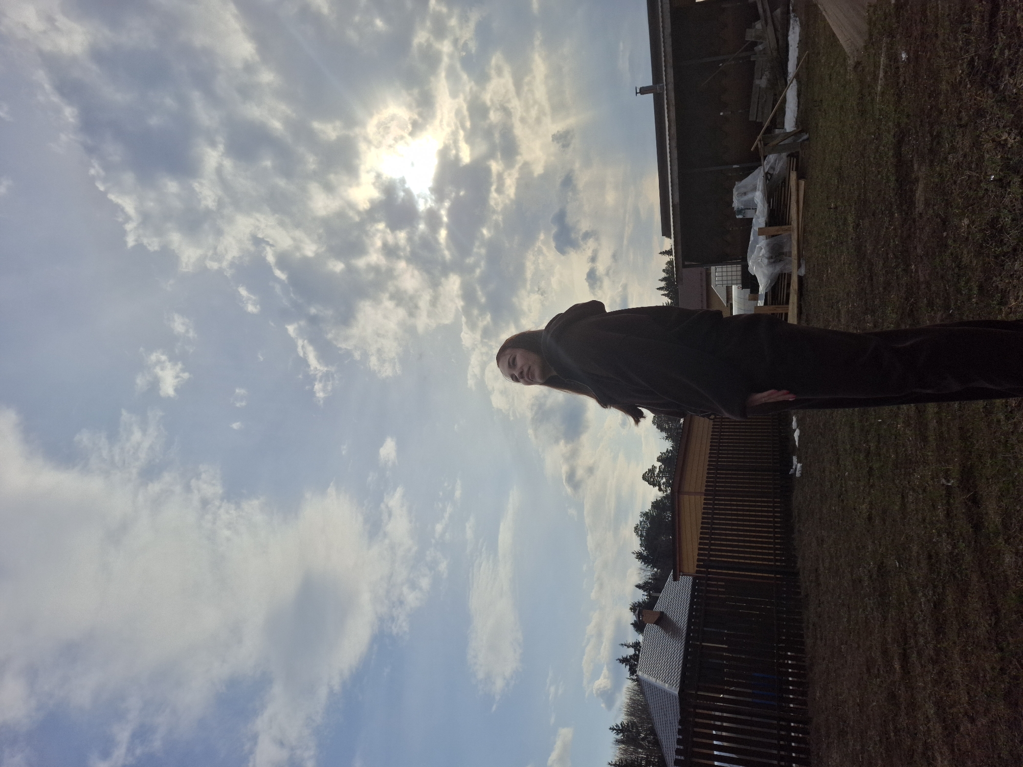 The image depicts a woman standing in a yard in Chaikovsky, Russia. The sky is cloudy with a visible sun. There are buildings, a fence, and grass visible in the background.