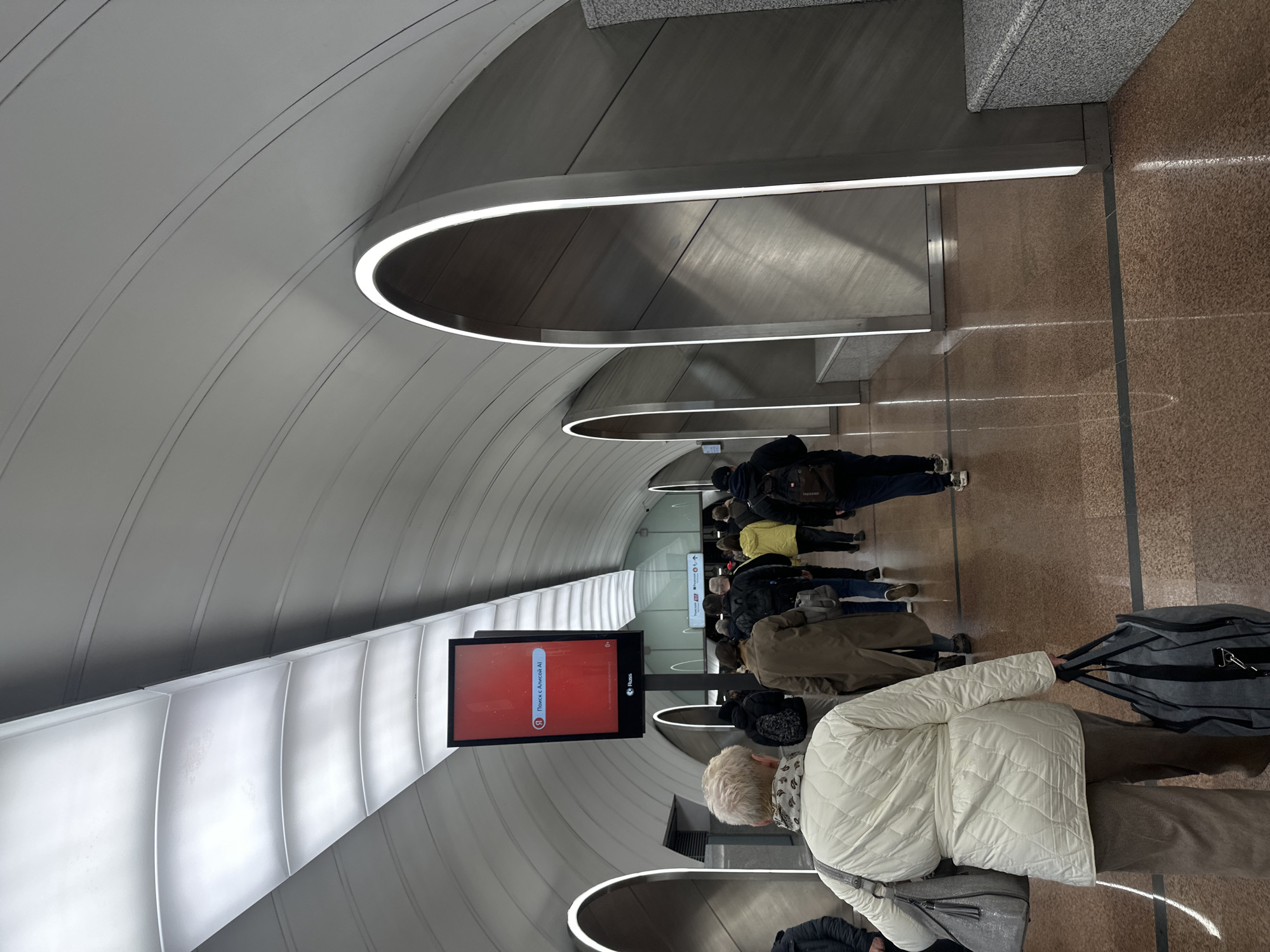 The image depicts a queue of people in what appears to be a modern, underground space, likely a subway station. The setting is suggested to be in Moscow, Russia based on the Russian language text visible on a digital display. The station has a futuristic design with a curved ceiling, integrated lighting, and tiled walls. A line of people is visible, presumably waiting for transportation. Most are dressed in winter clothing. A digital screen showing advertising or station information is present on the left. A few people have luggage indicating that they may be traveling.
