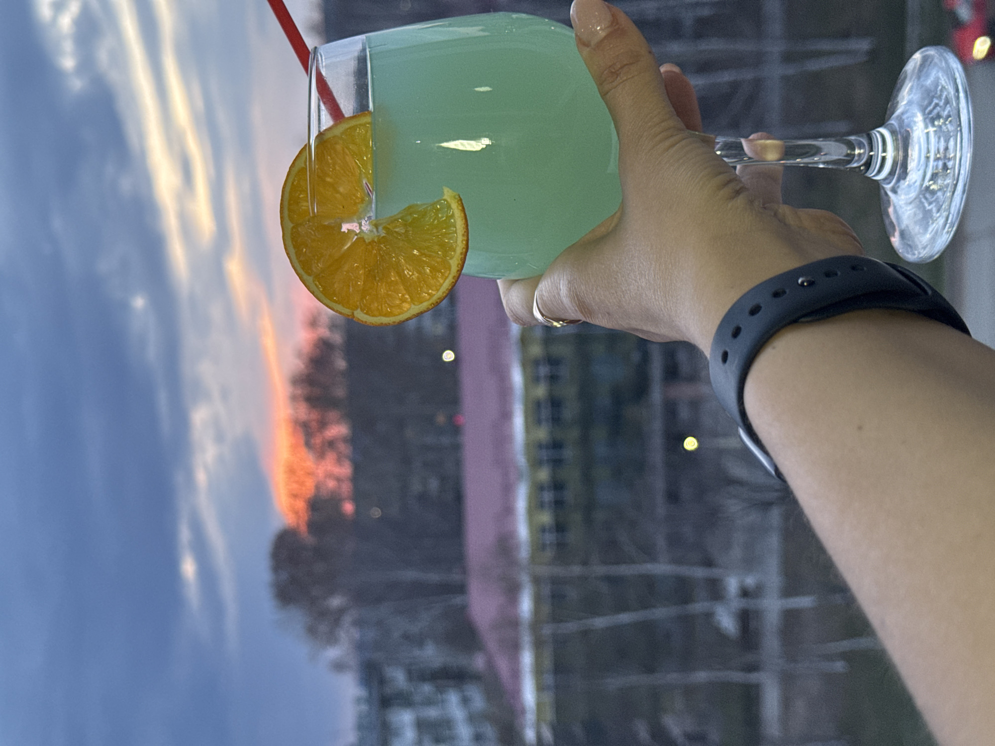 A vertical photograph captures a person's right hand holding a stemmed glass with a beverage against a sunset backdrop. The hand, with fair skin and manicured fingernails painted a nude color, features a silver ring on the ring finger and a dark gray smartwatch on the wrist.
The glass contains a pale blue-green drink and is garnished with a bright orange slice on its rim. A red straw is also visible in the glass.
The background, viewed through a window, shows an outdoor scene during sunset or dusk. The sky displays a gradient of colors, from blue at the top to vibrant orange, pink, and golden hues near the horizon, illuminated by the setting sun. Streaky clouds reflect these warm tones. Below the sky, blurred forms of distant buildings or structures and possibly trees are visible.
The image suggests a moment of relaxation, with the individual enjoying a drink while observing the scenic evening sky. The implied location is Chaikovsky, Russia. There is no legible text visible in the image.