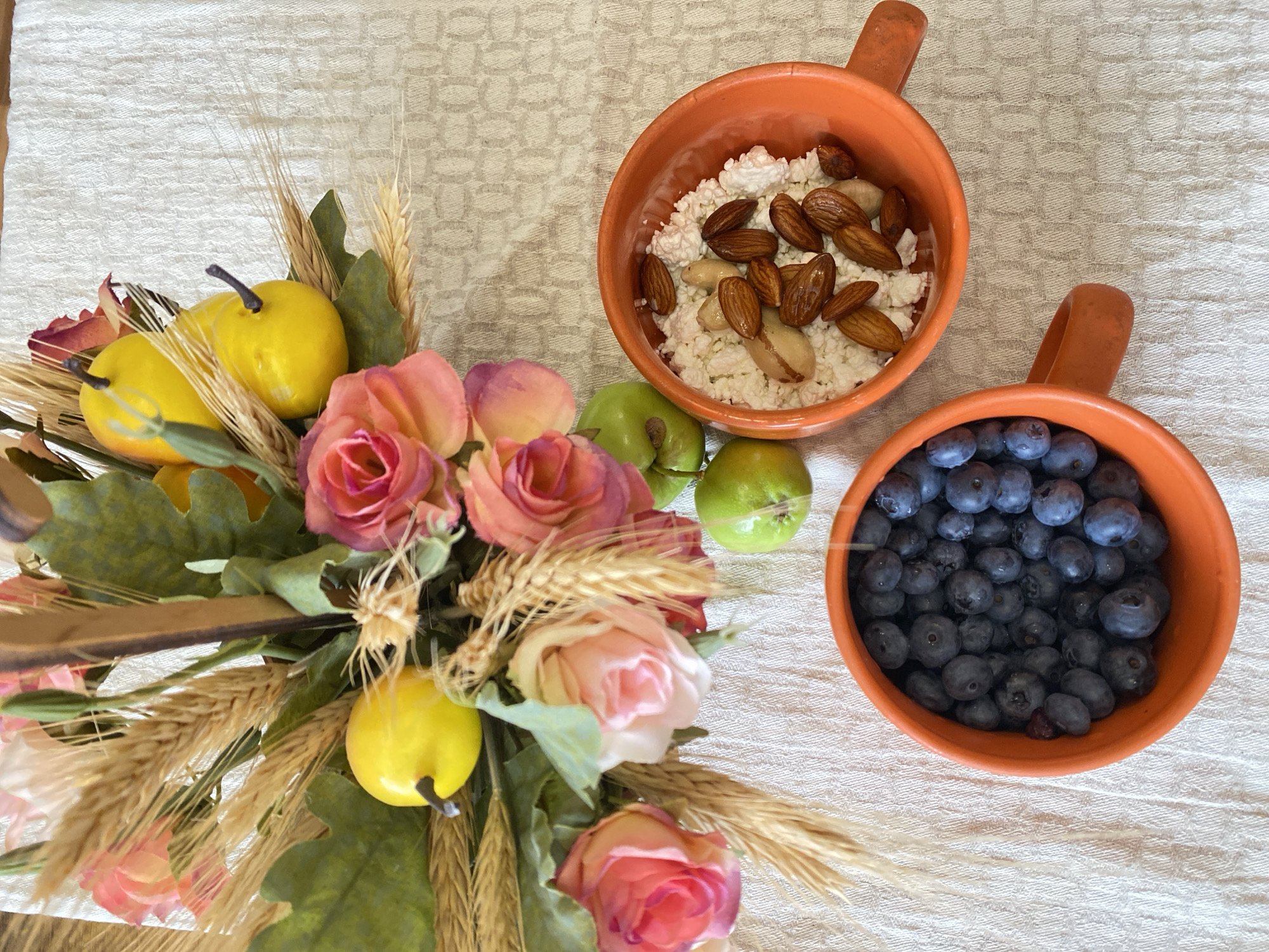 The image displays a still life arrangement on a textured white tablecloth. In the upper left quadrant, a decorative bouquet features artificial pink roses, green leaves, and wheat stalks. Interspersed within the bouquet are two bright yellow, pear-shaped decorative objects and two small, green apples are placed in front of the bouquet. To the right of the bouquet, two orange ceramic bowls are positioned on the tablecloth. The bowl on the right contains cottage cheese topped with almonds and what appear to be Brazil nuts. The bowl on the left is filled with fresh blueberries. The scene is brightly lit, with natural light casting subtle shadows. There are no people or animals visible in the image, and no specific landmarks or indicators of location within the city of Lida, Belarus are present. The context is that of a prepared breakfast or snack setting.