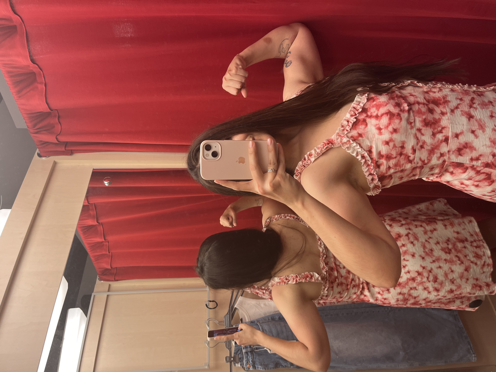 A young woman is posing for a mirror selfie in a fitting room. She is wearing a white and pink floral print dress with ruffled straps. She is flexing her biceps, looking at her reflection in the mirror. In the background, there are red curtains and clothes on a rack. The scene appears to be a retail store. There is no indication of the location being Yessentuki, Russia.