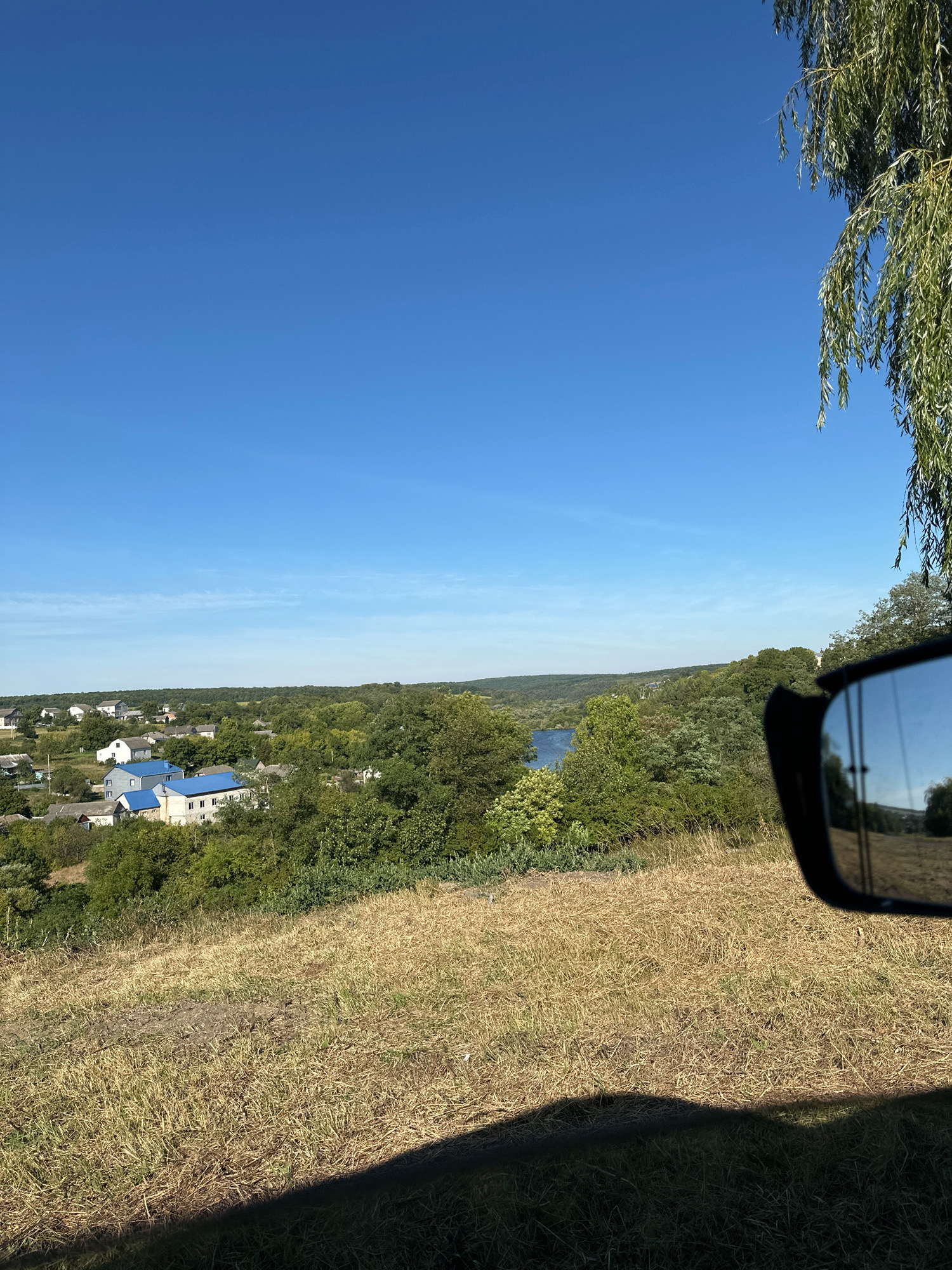 The image depicts a rural landscape under a clear blue sky, with a hint of a vehicle's side mirror visible on the right. In the middle ground, a village with houses featuring blue roofs is nestled among lush green trees. A body of water, possibly a river or lake, is visible beyond the village. The foreground is dominated by dry, golden grass, casting a shadow. The scene suggests a serene, sunlit day in a rural setting, potentially in the city of Makiv, Ukraine, given the context. The reflection in the side mirror shows a continuation of the landscape, including trees and a hazy sky.