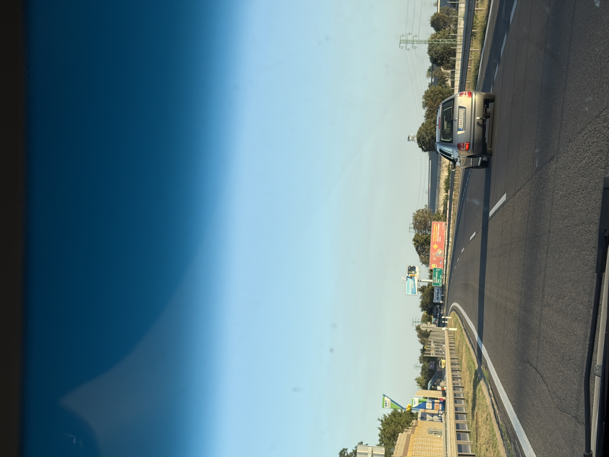 This is a vertical, slightly angled photograph of a highway in Vlčkovce, Slovakia. The scene is captured from within a vehicle, likely a car, looking forward. On the right side of the image is the asphalt road with white lane markings. In the middle lane, ahead and slightly to the left of the frame's center, a silver hatchback car with a license plate reading "BA 455DS" is traveling in the same direction.

The left side of the image is dominated by a gradient of blue sky, transitioning from a deep blue at the very edge to a lighter, almost white sky towards the center. On the right side of the highway, a grassy verge is separated from the road by a metal guardrail. Beyond the verge, there are trees and in the distance, power lines and pylons are visible. A few billboards are also present along the roadside, one with red and white signage, and another green sign with white text. A green triangular sign with white text, possibly a road sign, is visible closer to the foreground on the right. The time of day appears to be daytime, with clear, bright conditions suggesting good weather. There are no people visibly featured in the image. The overall impression is that of a typical highway journey.