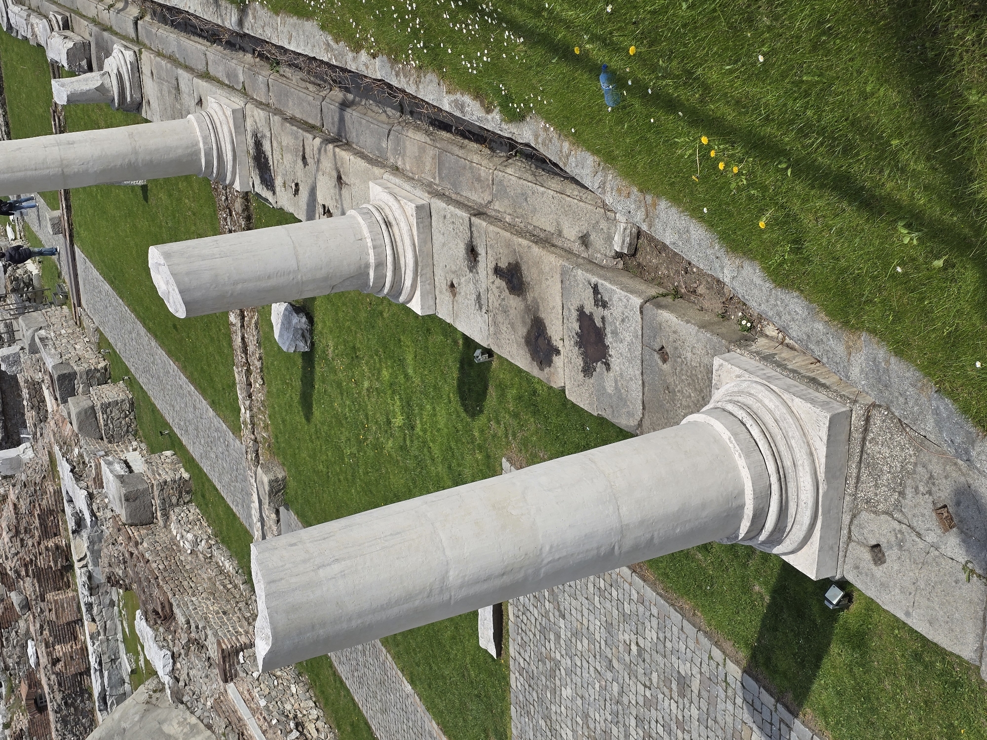 The image depicts the remnants of ancient architecture in an urban setting. Three weathered, white stone columns extend horizontally from a partially ruined wall. The columns appear incomplete, with fractured ends. Adjacent to the wall, a green grassy area is visible, interspersed with cobblestone pathways. A small number of people are present, observing the ruins. A blue plastic bottle is visible on the grass near the wall. The location is Unknown, Unknown.