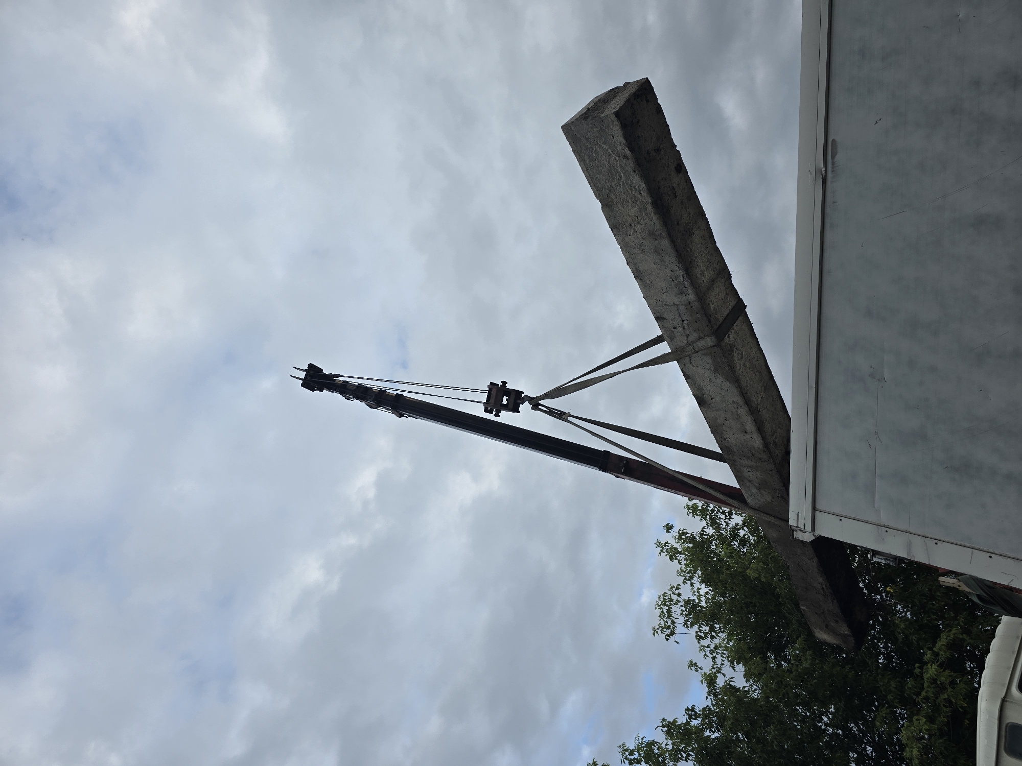 A crane is lifting a large, rectangular concrete beam, suspended by straps, over the side of a white truck. The truck's roof is visible, and a portion of a white vehicle is visible in the lower left corner. Trees with green foliage are to the left of the crane. The sky is overcast with gray clouds. The scene appears to be in Ермолаевский Затон, Russia, though no specific landmarks are identifiable.