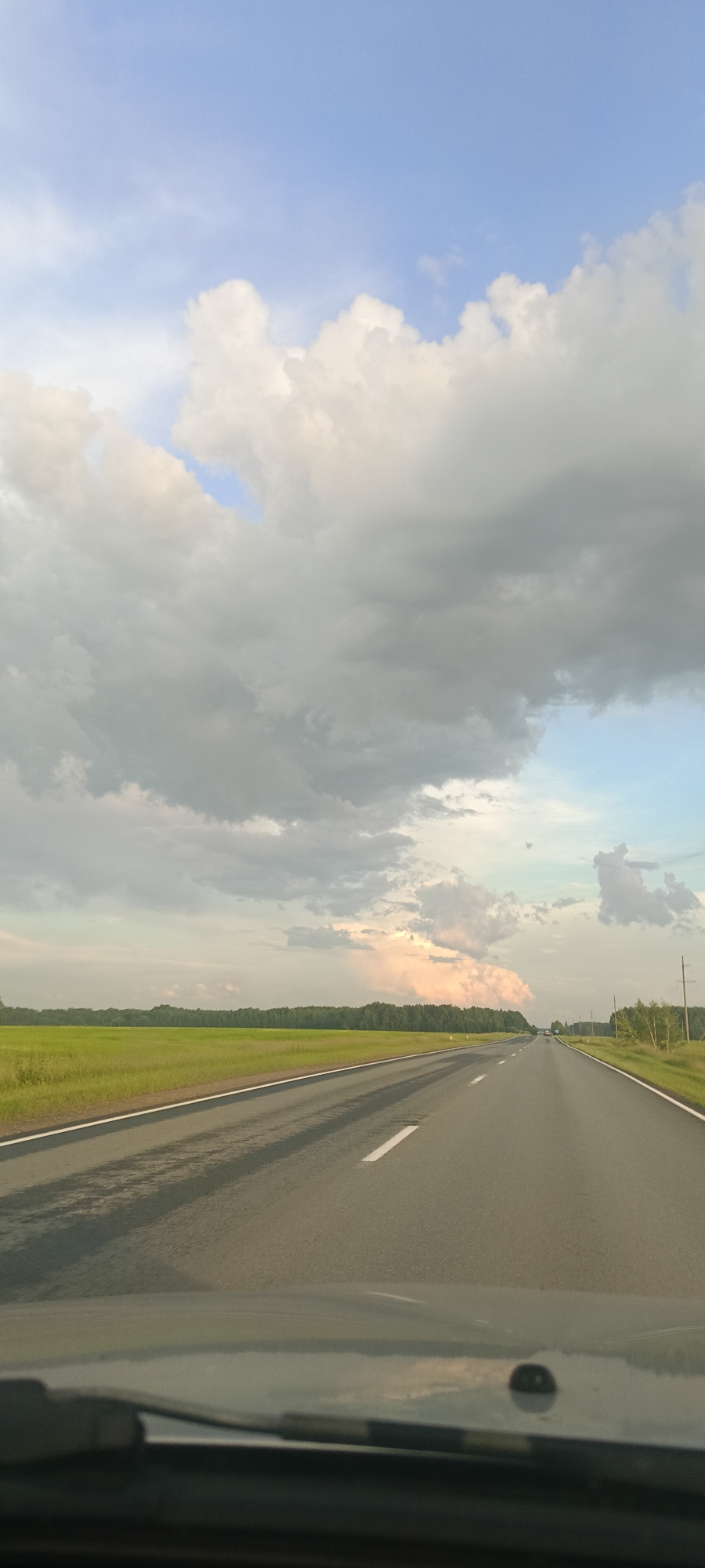 The image shows a view from inside a car driving on a two-lane road. The road stretches into the distance, bordered by a bright green field on the left and a line of trees on the right. The sky is filled with dramatic clouds, with patches of blue sky visible. The lighting suggests it is late afternoon or early evening, with the sun casting long shadows on the road. The overall mood is serene and expansive, evoking a sense of journey and open road. No people or text are visible in the image.
