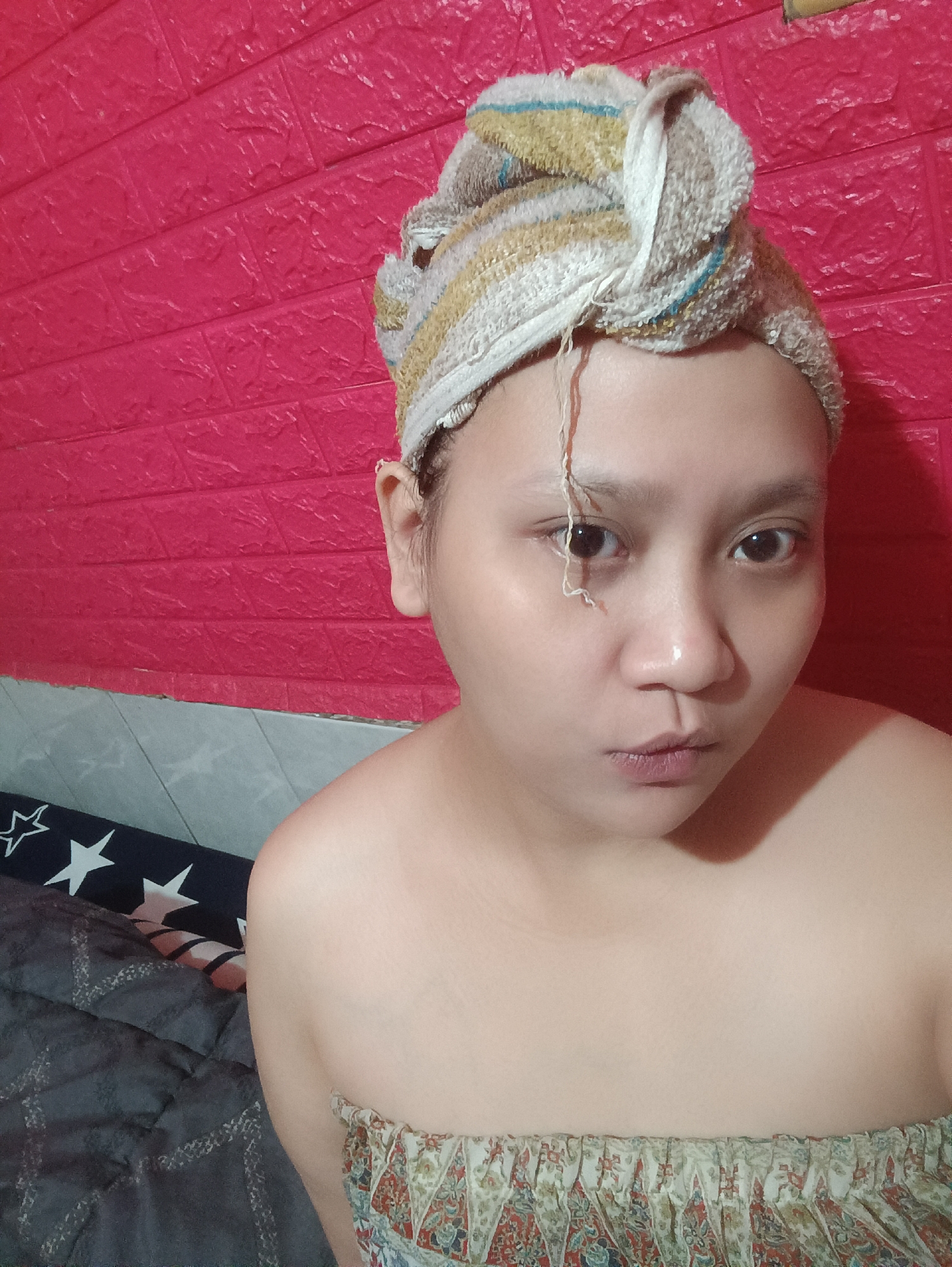 The media file is a selfie taken indoors, likely in a bedroom or bathroom, given the presence of a bed with a patterned duvet and a textured red wall, possibly wallpaper. A young person with a towel wrapped around their head is the sole subject. The towel is striped with yellow, white, and blue. A strand of what appears to be hair or a product residue is hanging down from the towel over their forehead and eye. The person's face is turned towards the camera, with a neutral or slightly pensive expression. Their eyes are open and looking directly at the viewer. They are wearing a strapless garment with a patterned fabric at the bust line. The background wall is a vibrant red with a raised, brick-like texture. To the left of the person, a portion of a dark duvet with white stars is visible, along with another patterned fabric, possibly bedding or upholstery, in muted colors. The lighting suggests an indoor setting, possibly during the day or evening. No text is visible in the image. The location context of Depok, Indonesia, provides a geographical marker for the image.