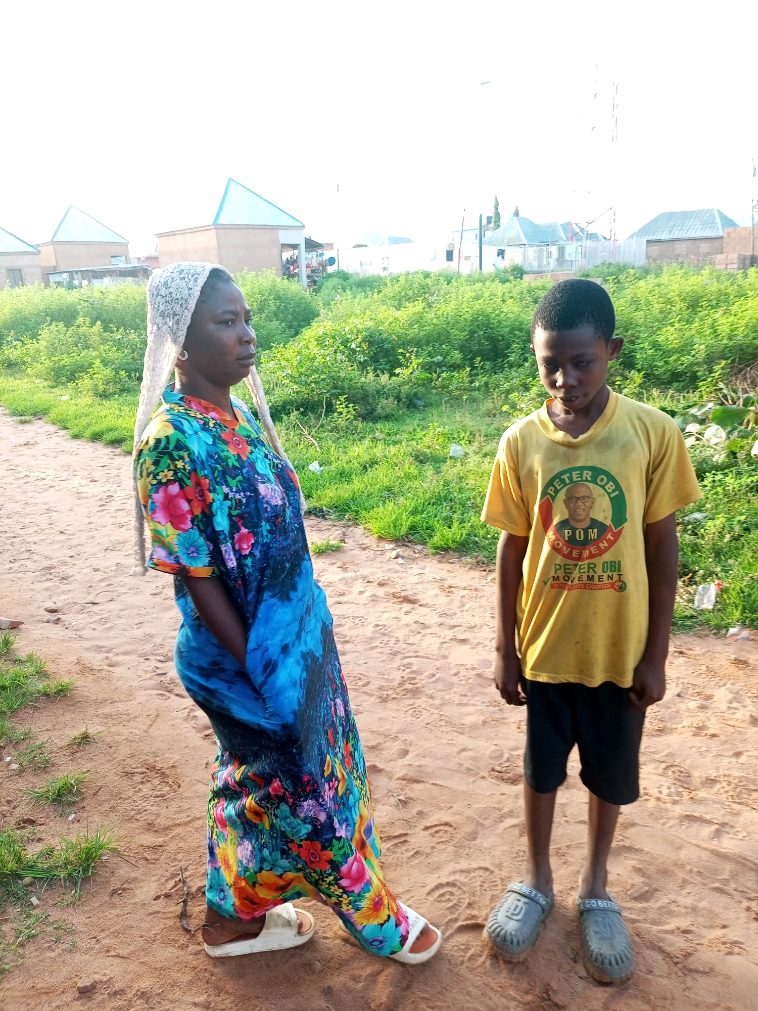 In Tempure, Nigeria, a young boy stands on a dirt path. He wears a yellow t-shirt with a graphic of a man's face and text that reads "PETER OBI MOVEMENT". He also wears black shorts and grey sandals. To his left, a woman stands wearing a headscarf and a brightly colored, floral patterned dress. She wears white sandals. Behind them, several buildings with pointed blue roofs are visible, surrounded by lush green foliage. The sky is bright and overcast.