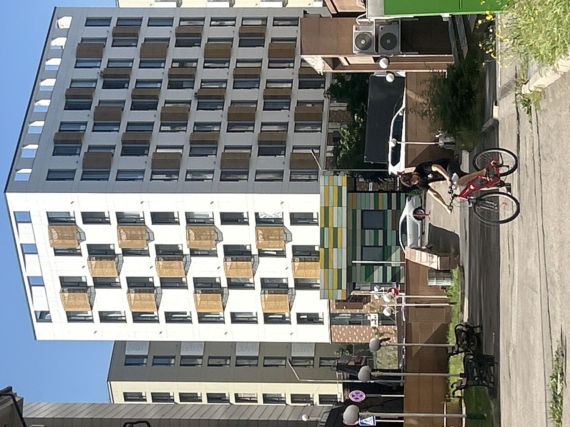 A man wearing a black t-shirt and black shorts is riding a red bicycle on a paved area. He is facing to the right of the frame and appears to be moving forward. Behind him, a modern white building with many windows and some decorative wooden elements can be seen. To the left of this building, another lighter-colored building with a similar facade is visible. In the foreground, there are benches and some greenery. The scene is outdoors, with a clear blue sky overhead. The overall impression is an urban setting, likely in Almaty, Kazakhstan, given the architectural style and the presence of modern residential buildings.