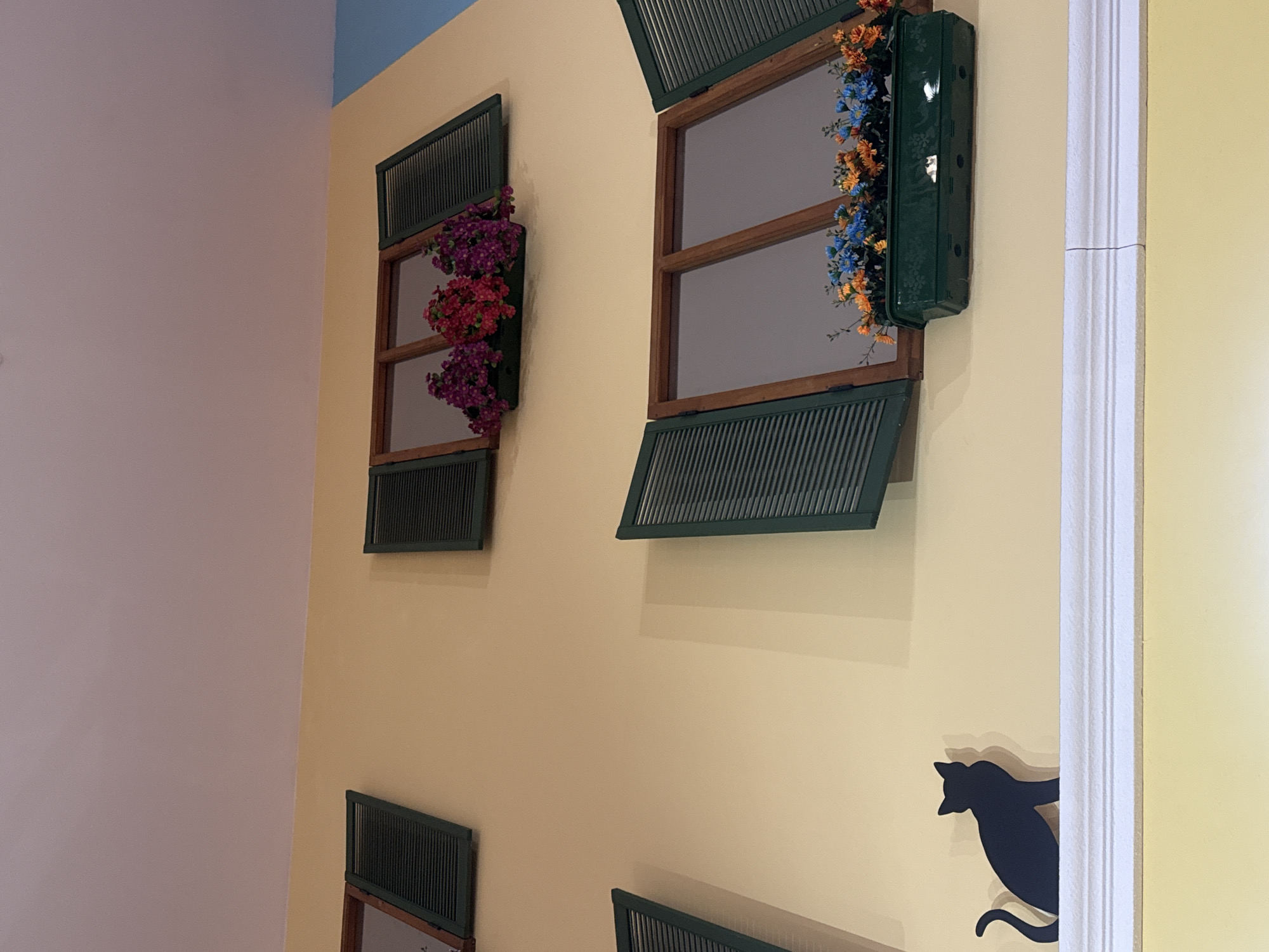 The image captures an interior scene, likely a room or hallway, with a distinct architectural style. The walls are painted in a warm, pale yellow, and the scene is illuminated by ambient light. The left side of the image reveals a section of a wall painted in a soft lavender hue.
Prominent in the composition are two faux window displays mounted on the yellow wall. These window installations are designed with dark green shutters, which are partially opened, revealing an artificial interior. The window on the left contains an arrangement of pink and purple artificial flowers, while the window on the right is adorned with a mix of orange and blue artificial flowers, along with a dark green planter box.
Below the window displays, there are additional, similar window elements, suggesting a pattern or theme in the room's decor.
Near the right edge of the image, a white architectural column or doorway is visible, creating a vertical line that adds depth to the scene. Adjacent to this column is a whimsical silhouette of a black cat, appearing as if it's peering around the corner.
Given the unique architectural details and the presence of stylized decorative elements, it is plausible that the image was taken in a creative or themed space within Yekaterinburg, Russia, although without additional information, it is difficult to precisely determine the exact location.