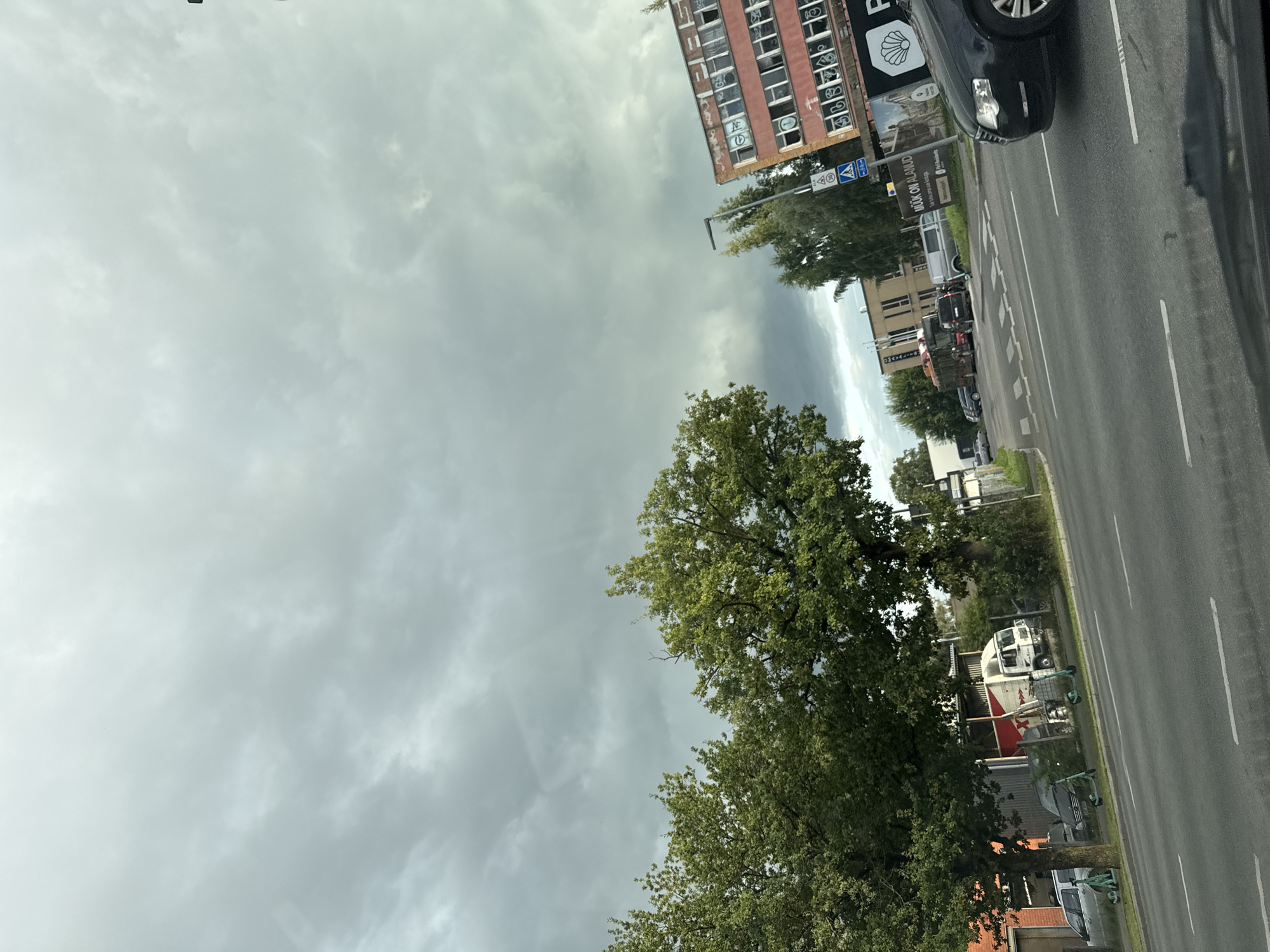The image shows a street scene in Tallinn, Estonia. The sky is overcast with dramatic, dark clouds. In the foreground, a road with white lane markings is visible. To the left, two large trees with green foliage are prominent. Behind the trees, there are parked vehicles, including a silver car, a dark-colored car, a white truck with a trailer, and two electric scooters on the sidewalk. Further down the street, there is a red building with multiple windows and graffiti. Adjacent to it is another building with a sign that reads "MÜÜK ON ALANUD." A street light and traffic signs are also present. A black car is driving in the right lane of the road.
