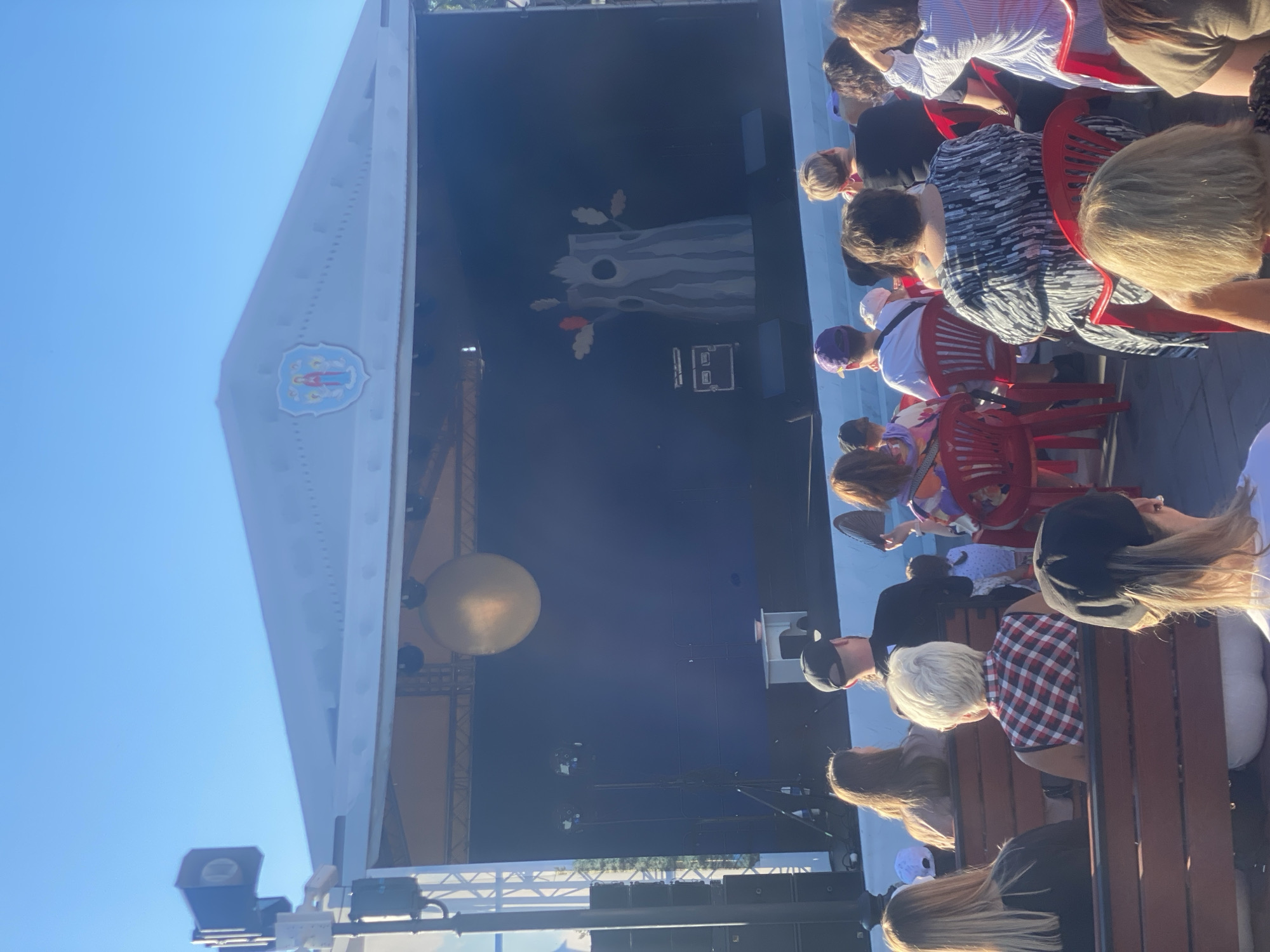 This image captures an outdoor stage performance, likely a puppet show or children's theater, in Minsk, Belarus. The stage is set against a clear blue sky, with a large, golden sphere suspended above. A backdrop depicts a stylized tree trunk with branches and leaves. In the foreground, a diverse audience of men, women, and children are seated on benches and red plastic chairs, facing the stage. Their attention is directed towards the performance. The overall atmosphere suggests a community event or festival, possibly in a park or public square.