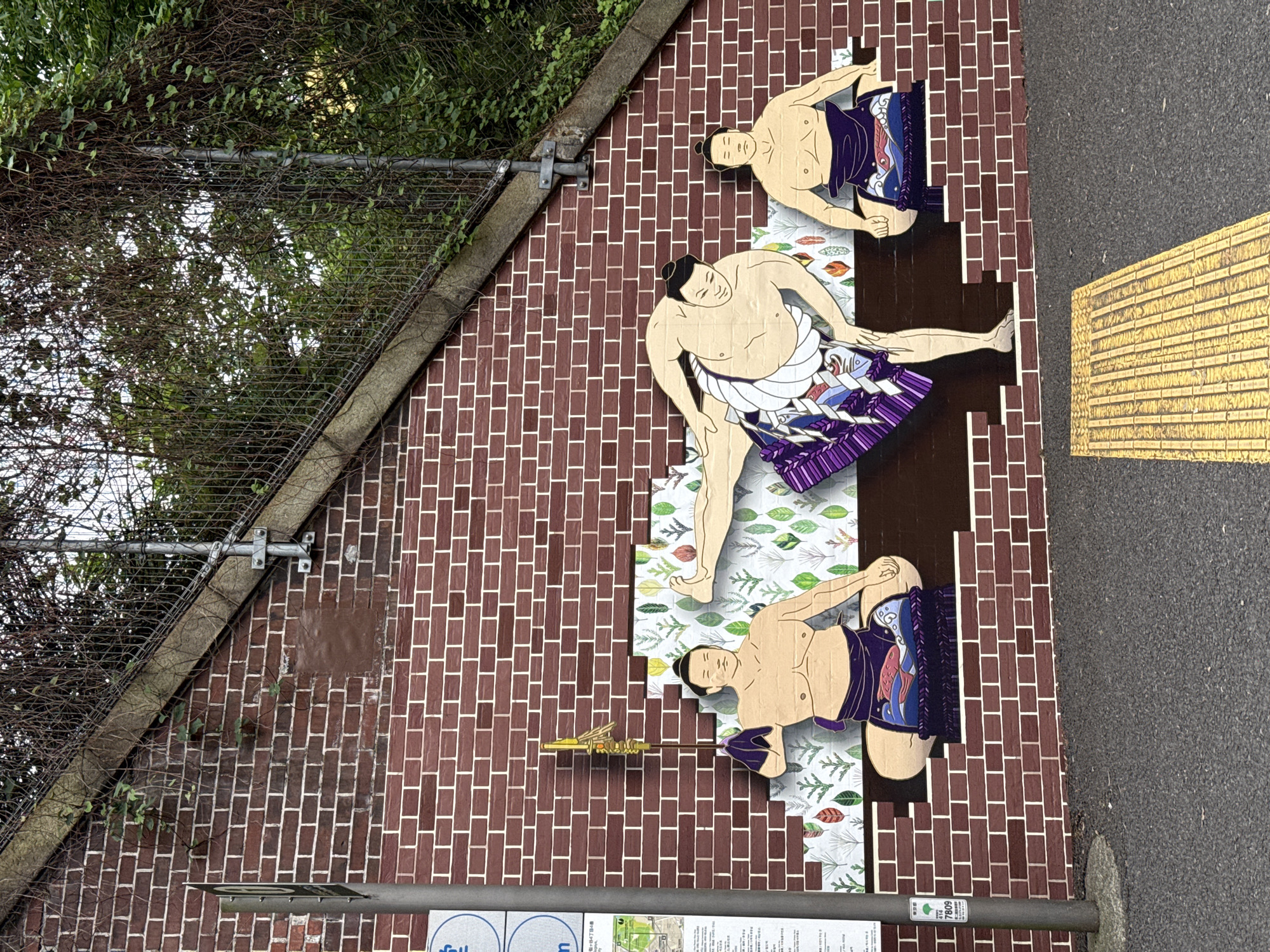 The image captures a street-level perspective of a brick wall mural in Shibuya, Japan, featuring three stylized sumo wrestlers depicted in various poses. The wrestlers are positioned against a background of brickwork, interspersed with patches of leaf patterns. The mural is located adjacent to a street, indicated by the presence of a paved sidewalk with yellow tactile paving, and an adjacent metal fence, partly obscured by foliage. A street sign is present at the bottom of the frame.
