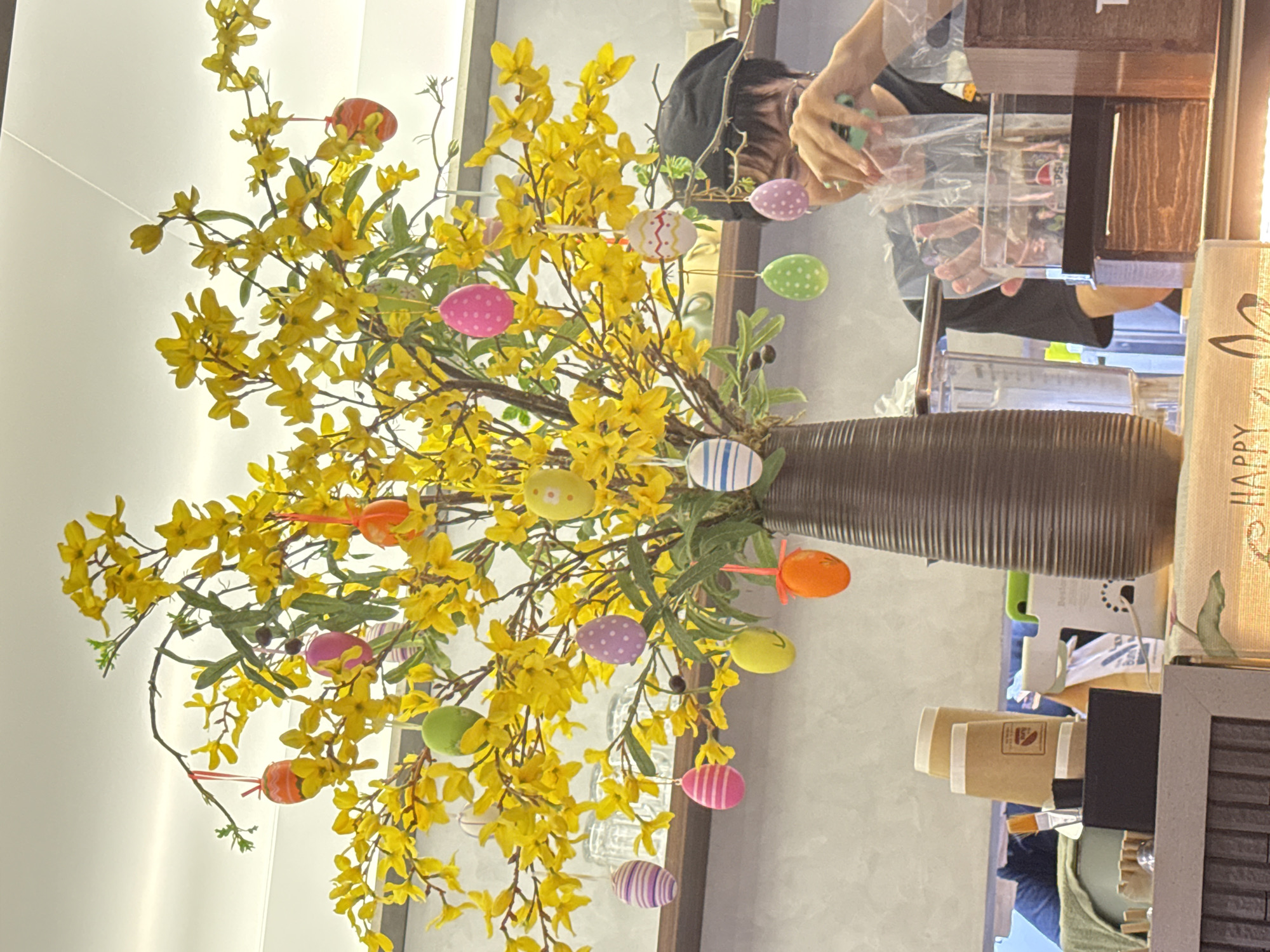 The image captures a scene with Easter decorations. A vase filled with blooming yellow flowers and adorned with colorful Easter egg ornaments takes center stage. In the background, an individual is visible, seemingly working behind a counter. The setting appears to be a commercial space, possibly a shop or café. Although I am unable to determine the location to be Đà Nẵng, Vietnam from this image.