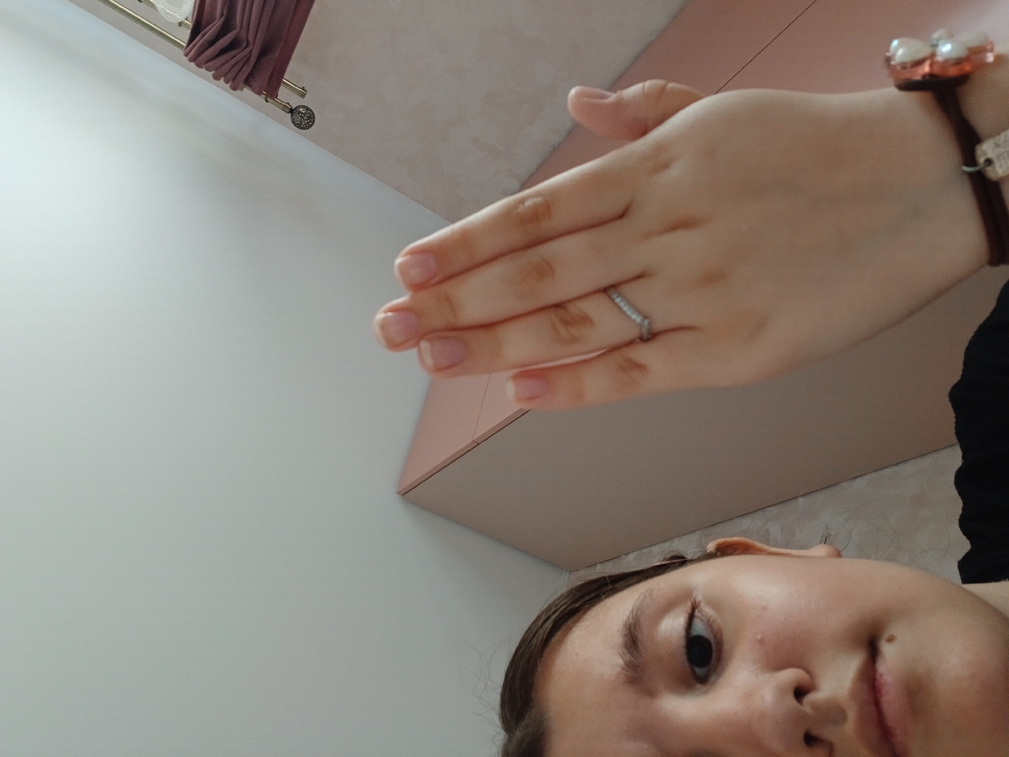 A close-up, slightly angled shot shows a person's face and hand. The face is in the lower right corner, showing the right eye, nose, and part of the mouth. The hand is held above the face, palm down, with a ring on the ring finger and a bracelet on the wrist. The background consists of a light-colored wall on the left, a pinkish surface behind the hand, and what appears to be part of a curtain rod with draped fabric in the upper left. The location is unspecified other than potentially being in Марковский, Russia.