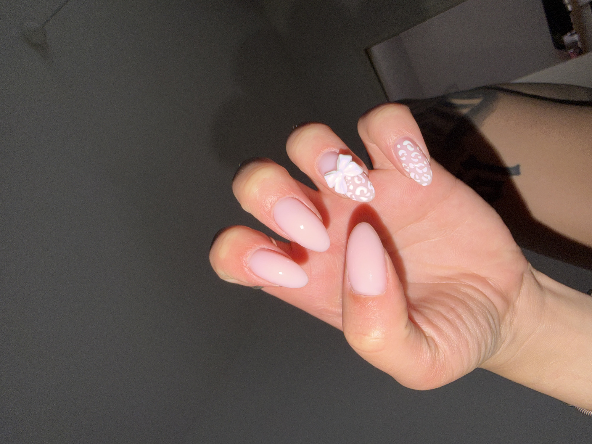 The image presents a close-up of a human left hand, held with the palm facing the viewer and fingers gently curled. The skin tone is fair to medium, with visible creases on the knuckles and wrist. All visible fingernails are almond-shaped and coated in a glossy, light pink polish. The ring finger's nail features a prominent, three-dimensional white bow embellishment. The pinky finger's nail is adorned with a pattern of white, irregular spots resembling leopard print, applied over the pink base. The thumb is partially visible on the left side of the frame. The background is dark and out of focus, appearing as a mottled dark grey to black, indicative of an indoor setting with limited illumination. In the upper right quadrant, a faint, blurred white object, possibly a smoke detector or ceiling fixture, is discernible. The lower right background contains a heavily blurred area consistent with an arm or shoulder, which appears to have a tattoo, and indistinct architectural elements of a room. The specific location within the city of Plovdiv, Bulgaria, cannot be determined from the visual information provided.